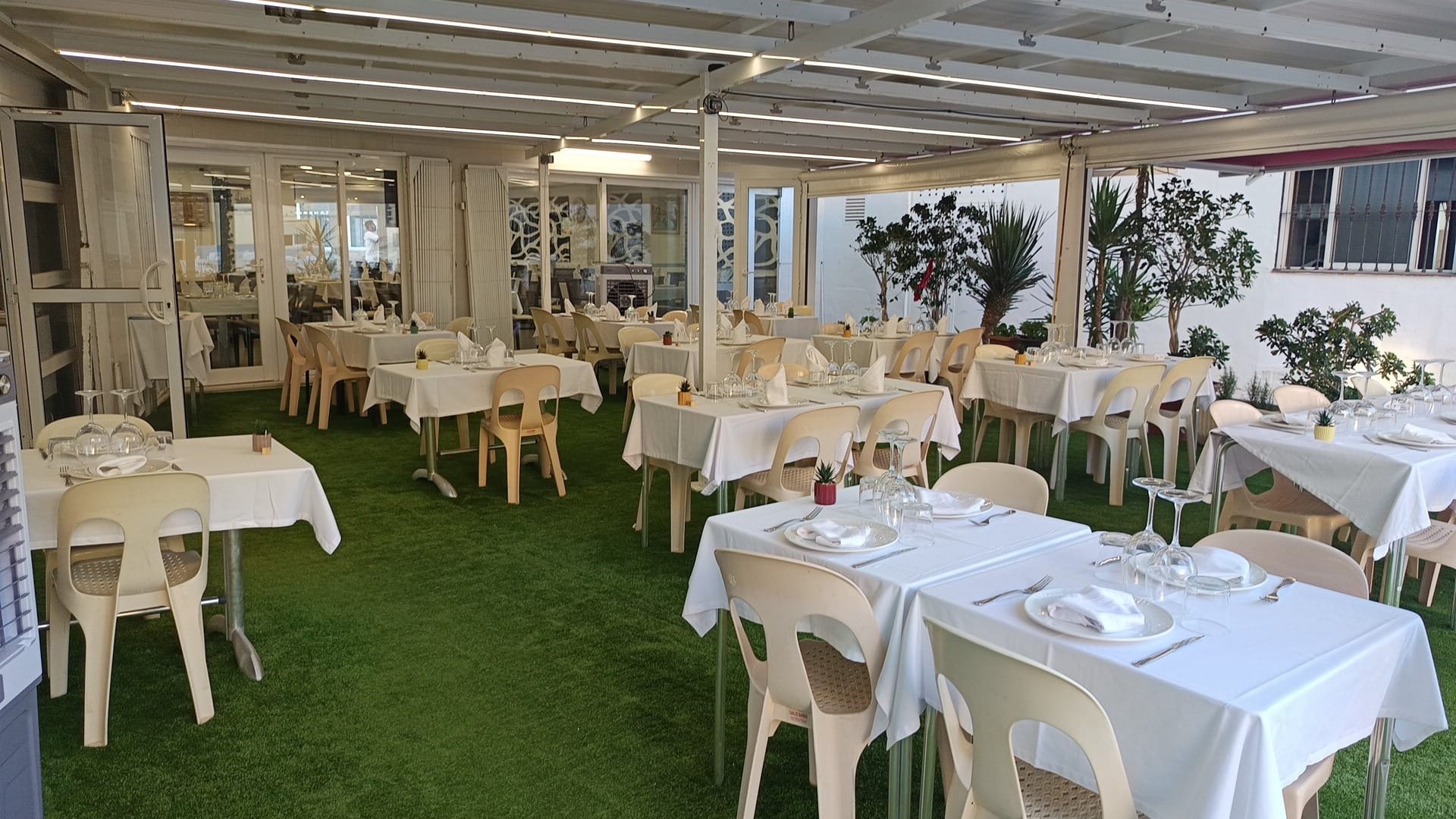 Comedor al aire libre con mesas y sillas, decorado con manteles blancos y plantas en macetas, ambiente luminoso y acogedor.