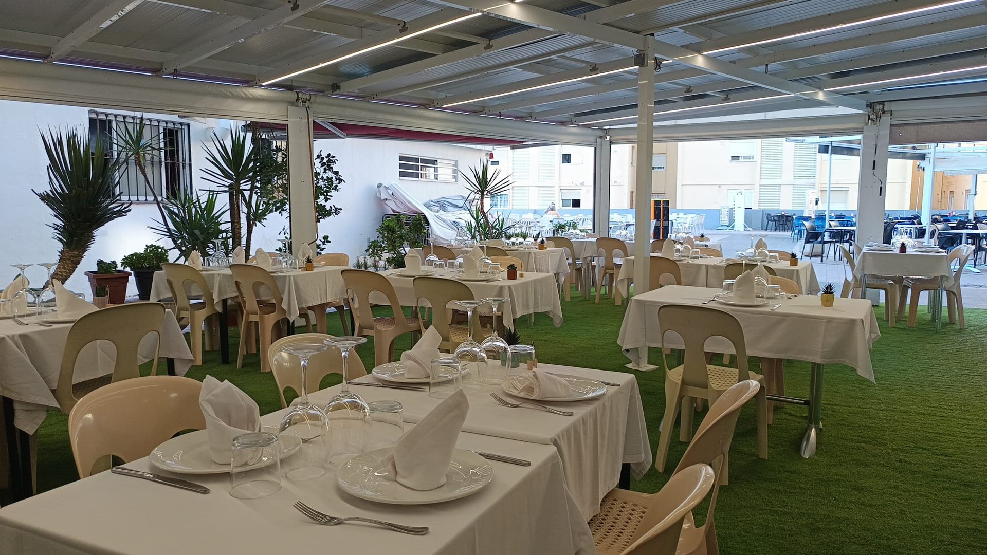 Comedor al aire libre con mesas vestidas con manteles blancos, cristalería y servilletas, decorado con plantas y césped artificial, en un espacio cubierto y techado.