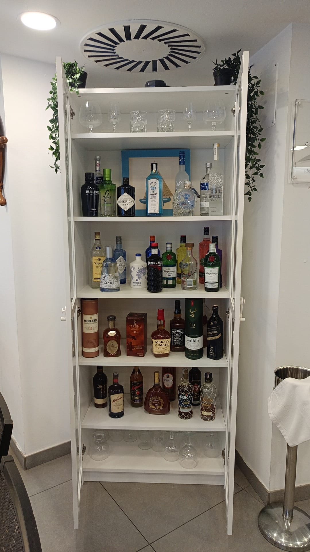 Vitrina blanca con varias botellas de licor y vasos en diferentes niveles, decorada con plantas en la parte superior y fondo blanco.