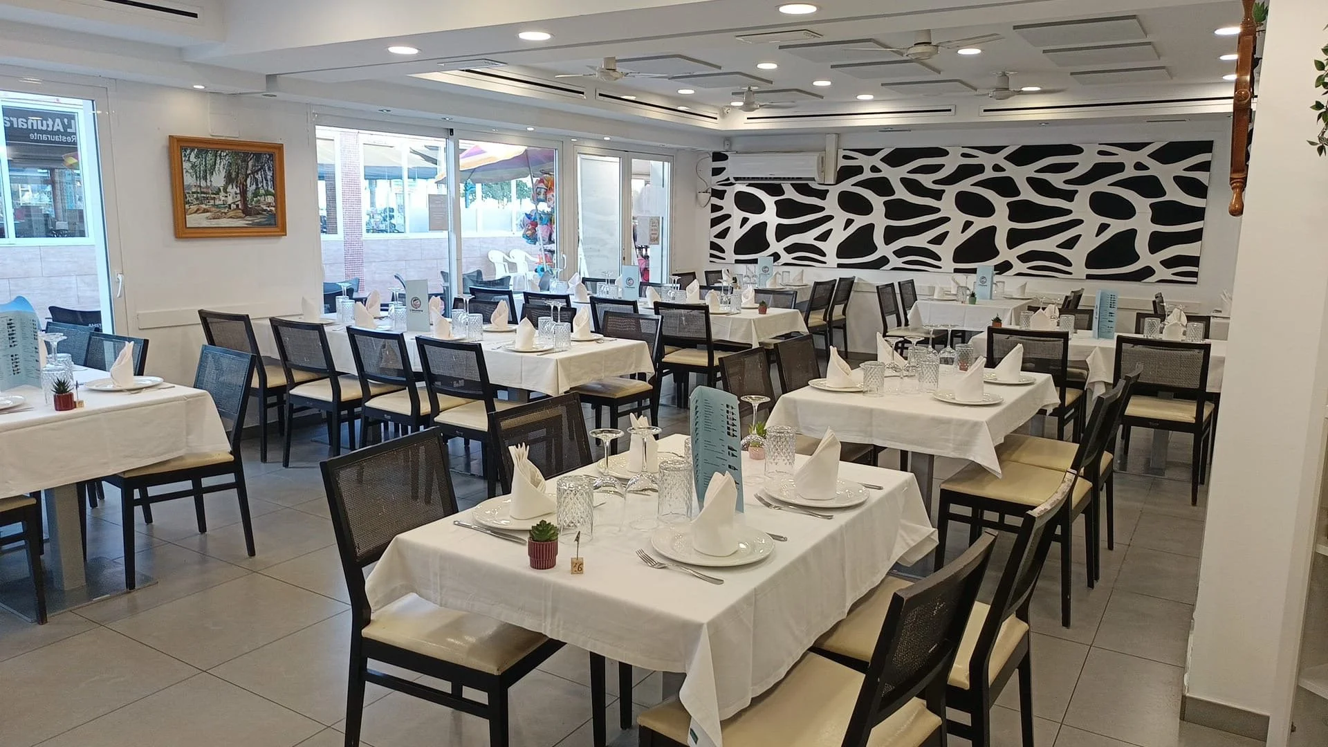 Interior de un restaurante con mesas arregladas con manteles blancos, cubiertos, copas y servilletas dobladas. Ventanas grandes permiten la entrada de luz natural, y hay decoración con cuadros y plantas.