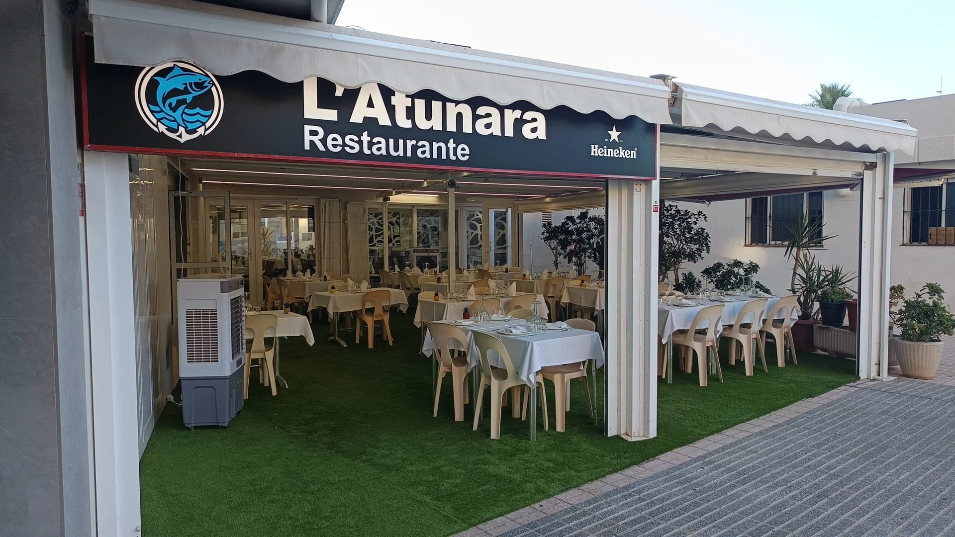 Fachada de un restaurante llamado 'L'Atunara', con área de comedor al aire libre con mesas vestidas con manteles blancos y sillas de plástico beige, decorado con plantas y cortinas de malla, con cartel de marca de cerveza Heineken.
