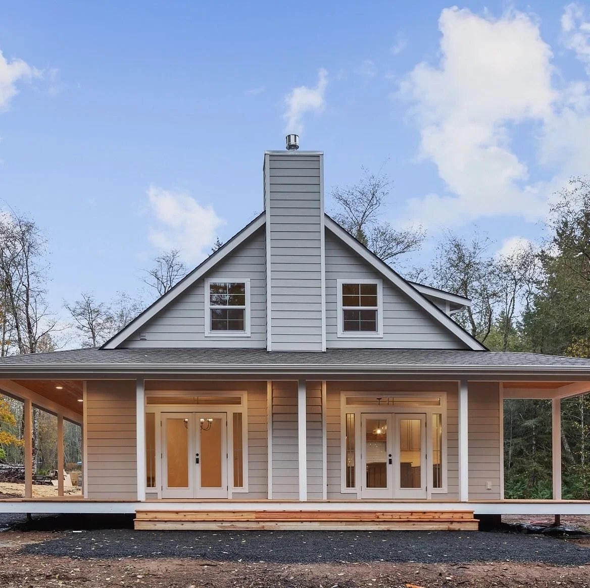 A modern two-story house with gray siding and a pitched roof, surrounded by trees, with a front porch and sliding glass doors.