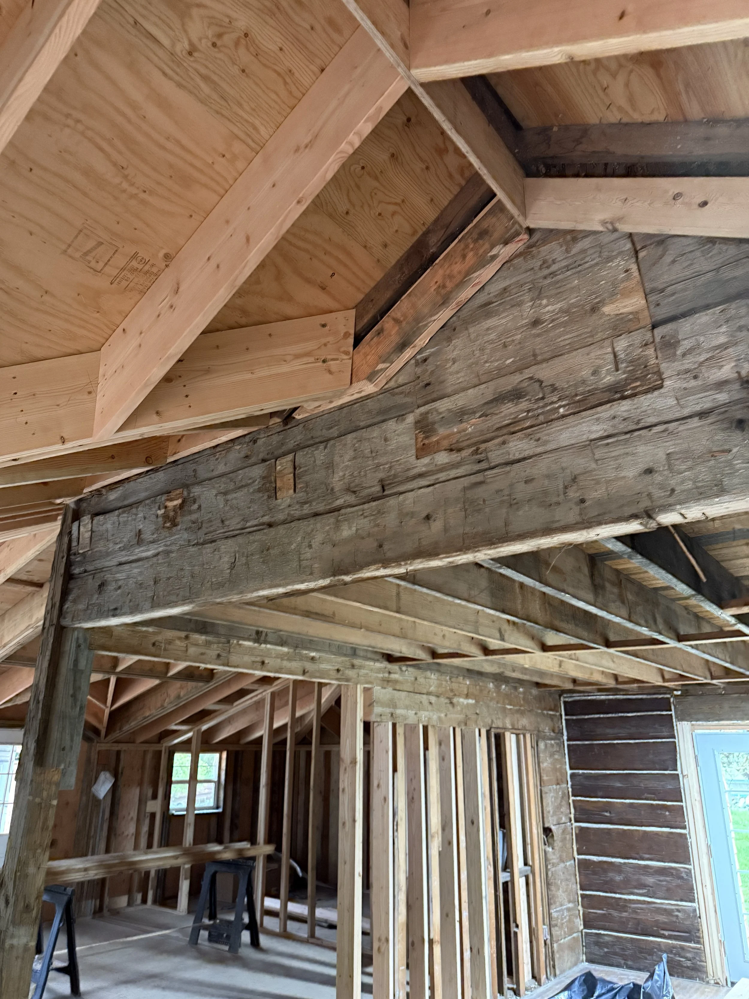 The interior of a building under construction with exposed wooden framing, beams, and wall studs, showing an unfinished ceiling and a partially completed wall with a window.