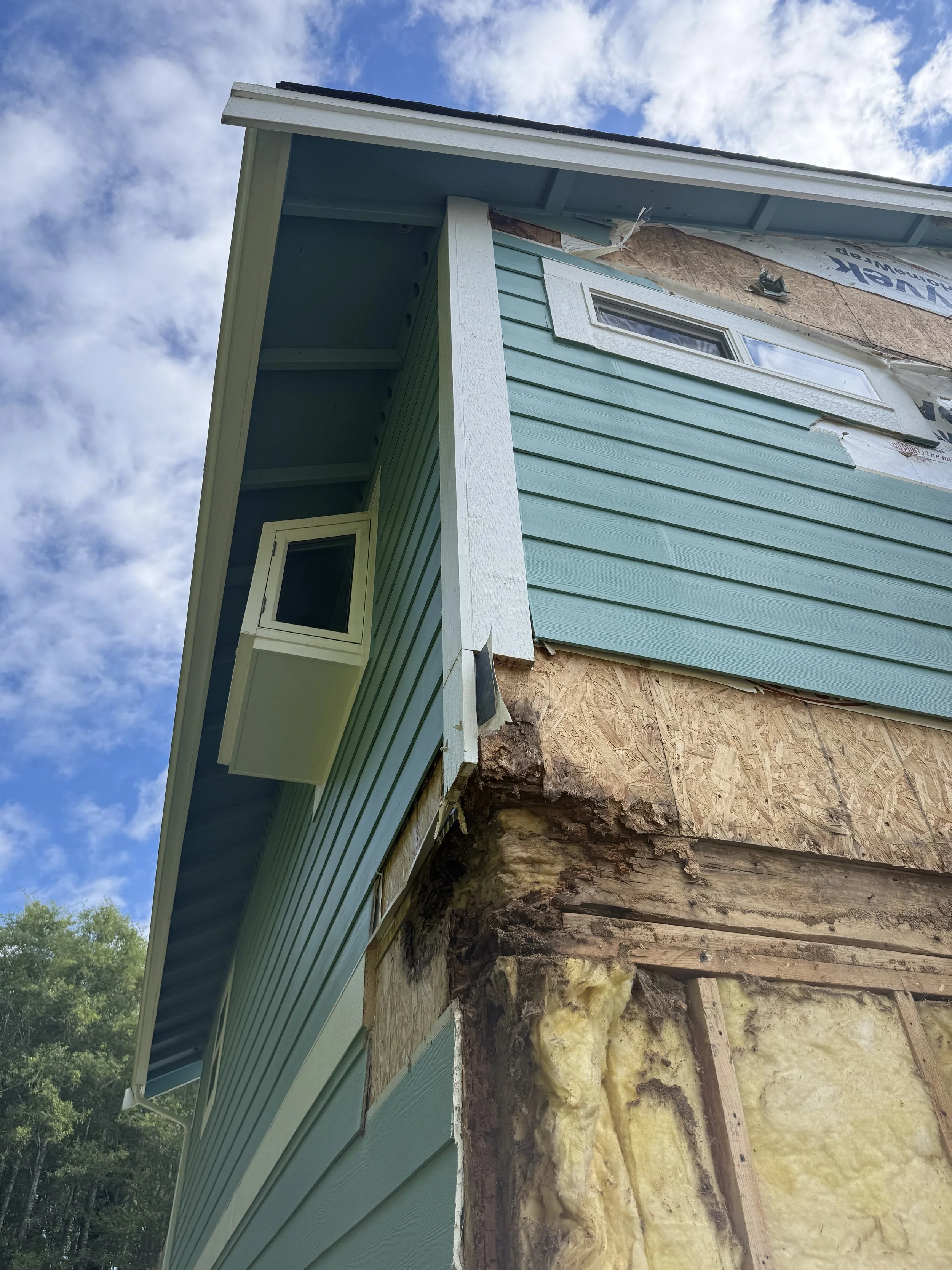 Side view of a house under construction, with blue siding and the weatherboard partially removed, revealing underlying insulation and wooden beams, against a background of a blue sky with clouds and some green trees.
