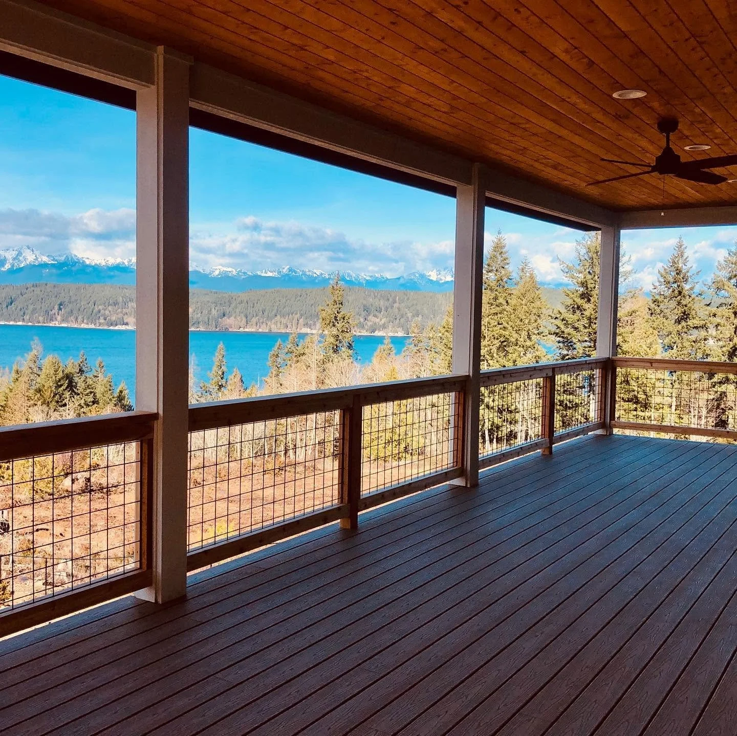 View of a balcony with wooden flooring and railing, overlooking a landscape with a lake, pine trees, and snow-capped mountains in the distance under a partly cloudy sky.