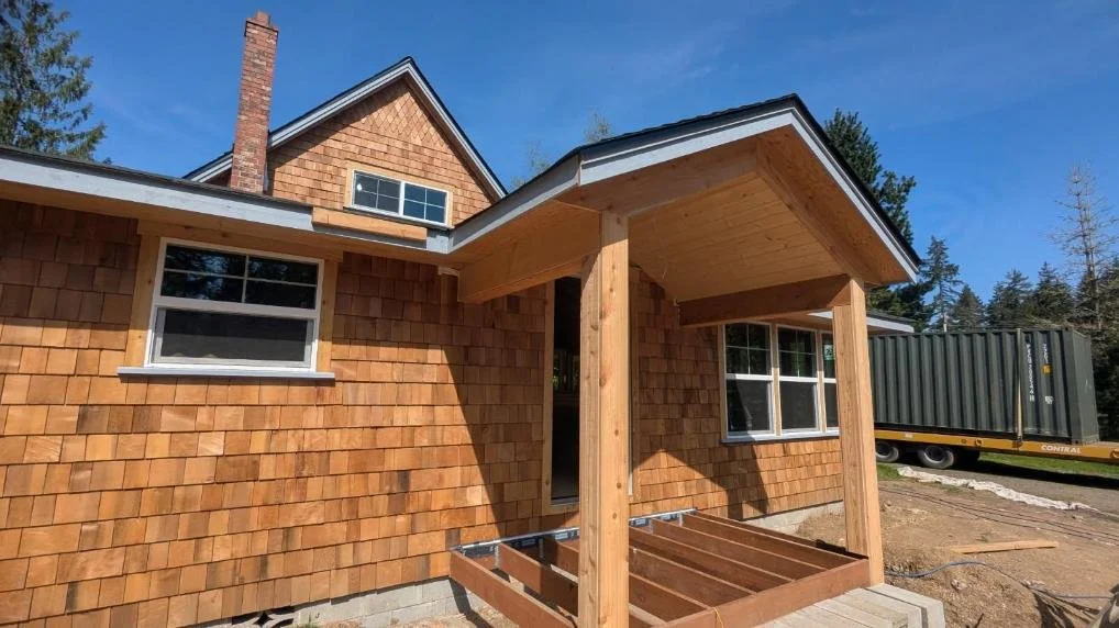 Construction of a new house with wooden shingles on the exterior, featuring a small porch with a wooden frame, and a shipping container in the background under a clear blue sky.