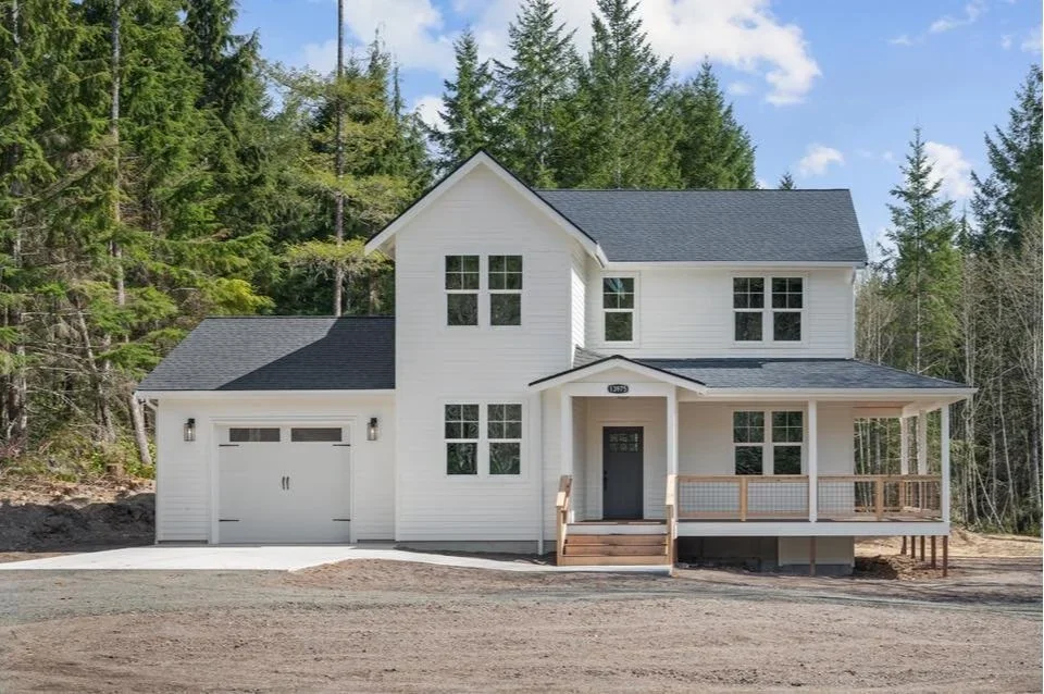 New two-story white house with a black roof, front porch, and attached garage, surrounded by trees under a partly cloudy sky.