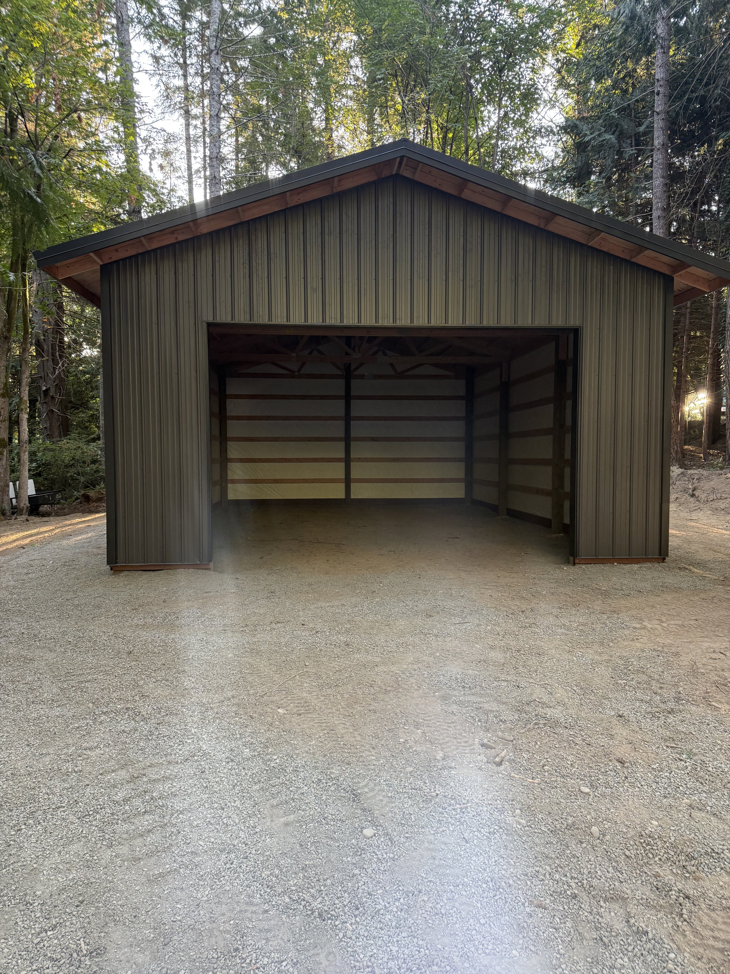A metal garage or shed in a forested area with trees behind and a gravel ground in front.