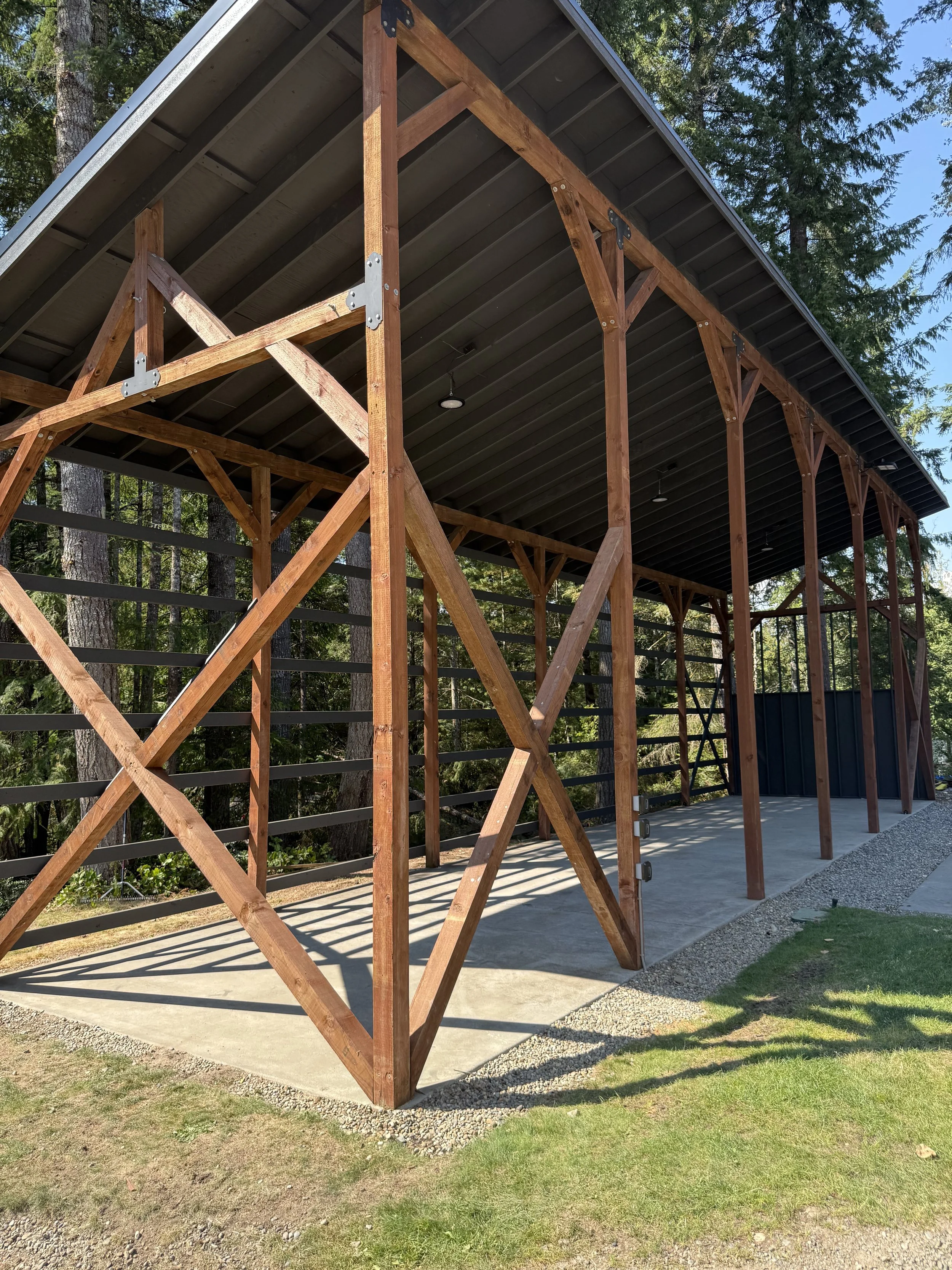 Side view of an outdoor building under construction with wooden framing, a concrete floor, and a metal roof, surrounded by trees and grass.