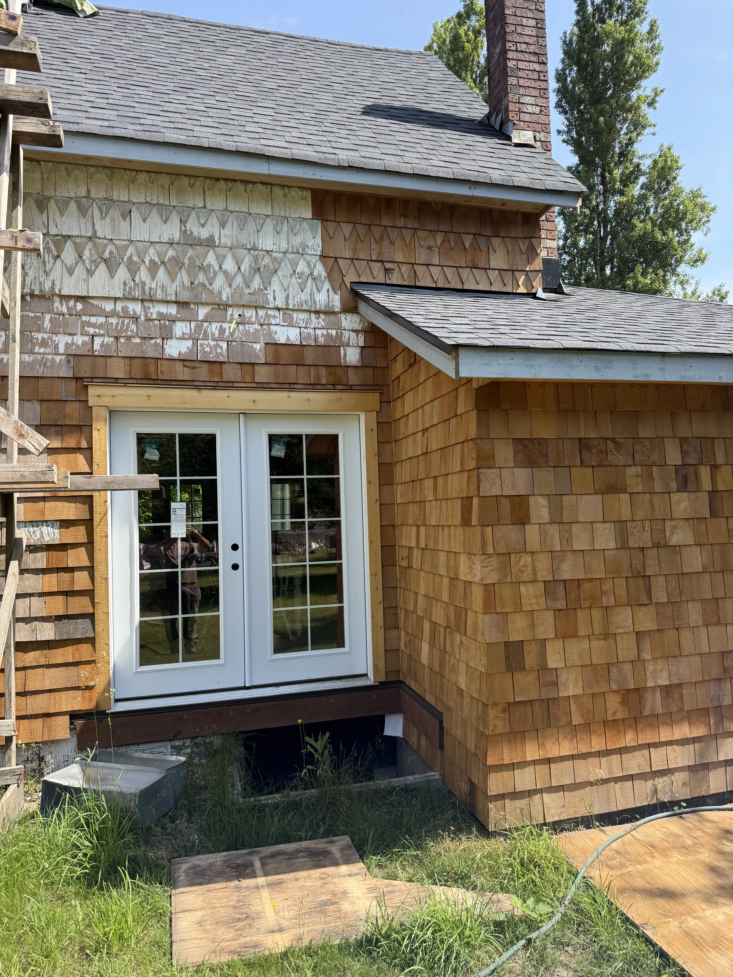 The back of a house under construction with a small porch and a French door. The house has wooden shingles and a mix of weathered and new wood siding. There is green grass and construction materials on the ground, with a tall brick chimney visible on