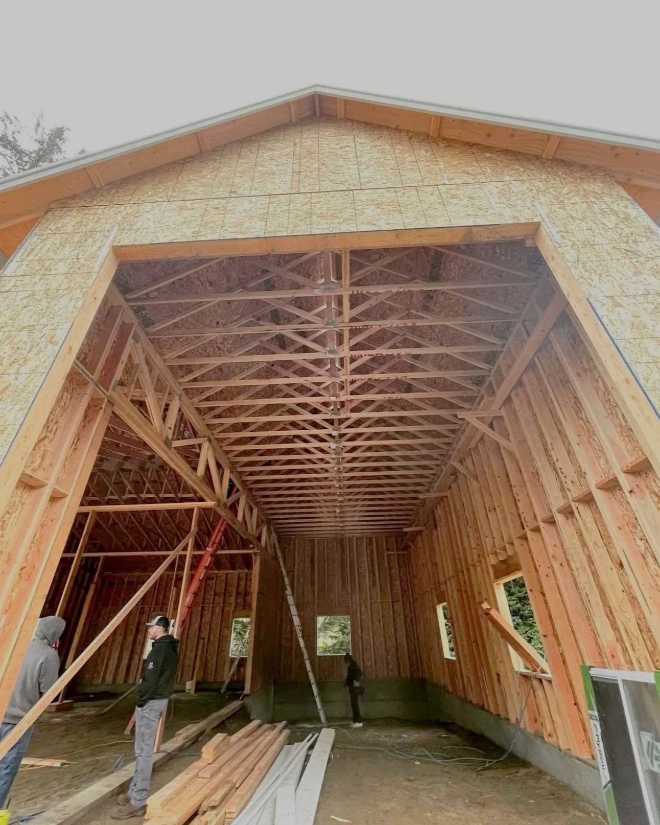 Interior of a building under construction with exposed wooden framing and roof trusses, no walls or roof yet, and three workers present.