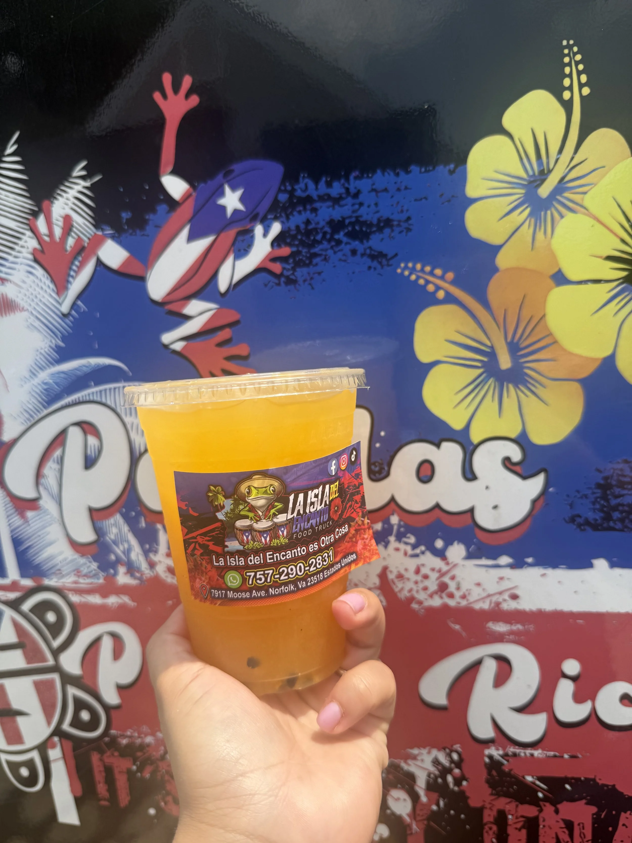 Close-up of a hand holding a yellow drink cup with a colorful label featuring a frog cartoon, a phone number, and addresses, against a background with vibrant floral and splatter decorations.