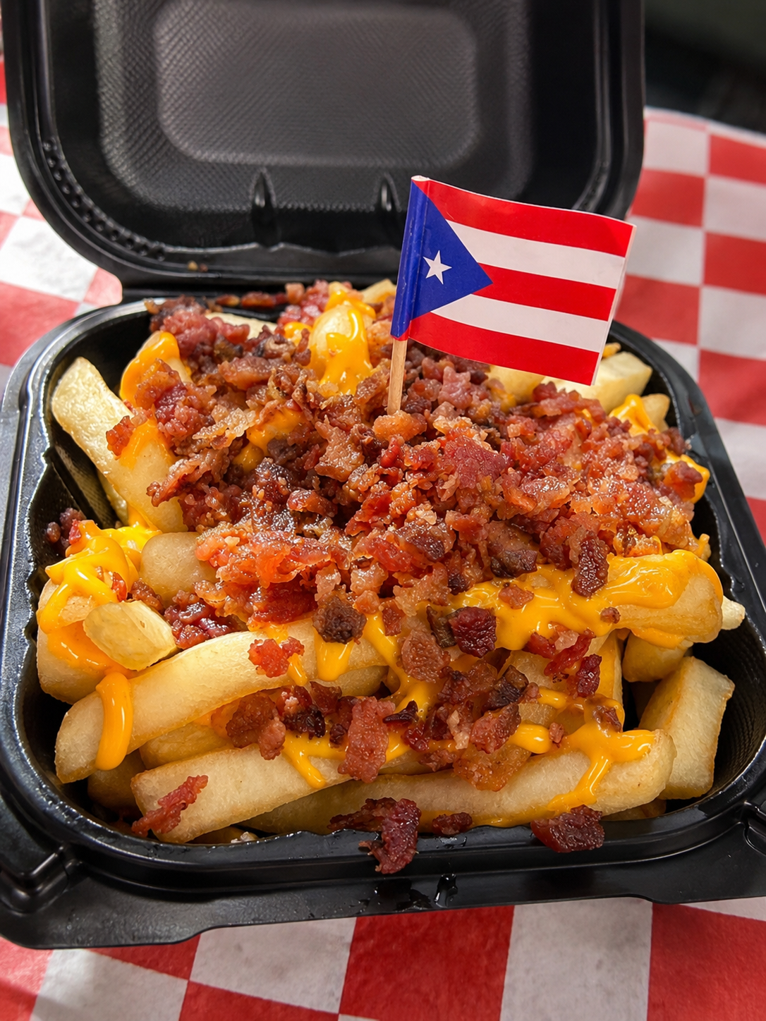 Container of loaded French fries topped with shredded cheese, bacon bits, and chopped bacon pieces, with a small Puerto Rican flag on top, sitting on a red and white checkered tablecloth.