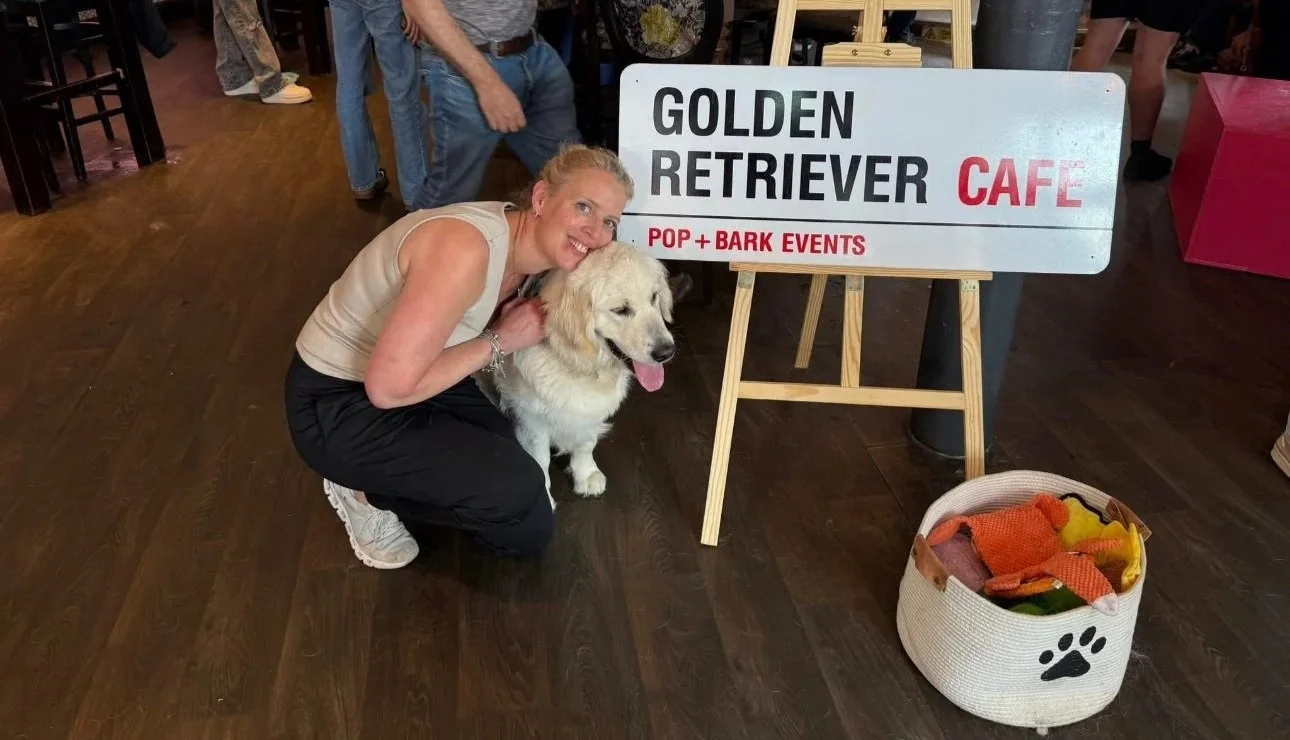 A woman kneeling next to a golden retriever inside a cafe, with a sign that reads 'Golden Retriever Cafe' and a basket of dog toys nearby.