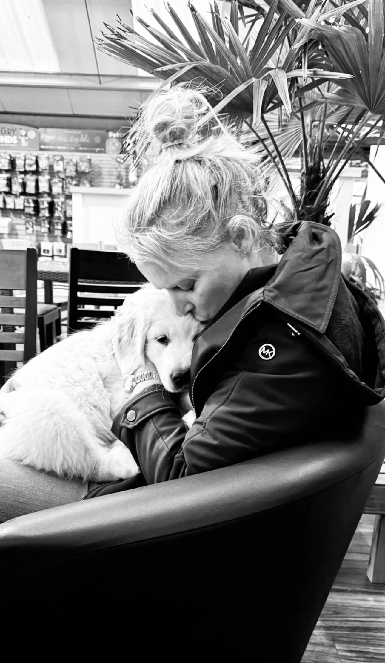A woman with light-colored hair in a ponytail is sitting on a bench, hugging a fluffy puppy. The puppy's head is resting against her chest, and she is gently pressing it with her face. There is a large potted plant with broad leaves behind them. The setting appears to be indoors, possibly in a store or a café, with shelves and signs visible in the background.