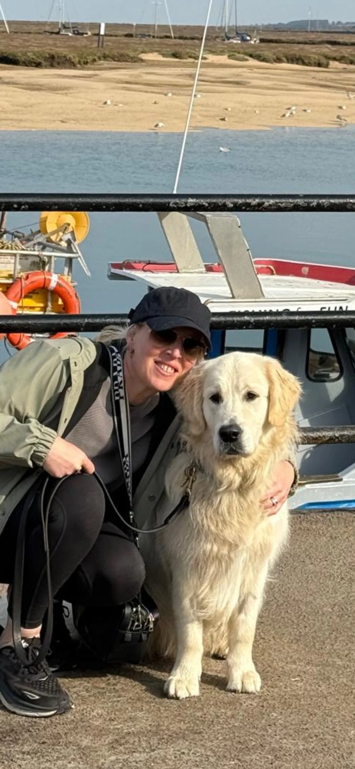 A woman with sunglasses and a black hat crouching beside a golden retriever puppy on a dock near a boat and water, with a shoreline and boats in the background.