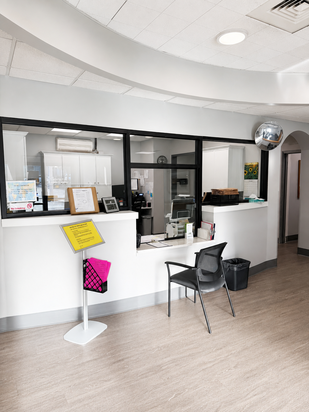Empty reception area with a black chair, a trash can, and a white counter with documents and office supplies, in a medical or office setting.
