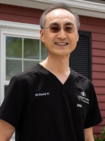 A smiling man wearing glasses and a black medical scrub top standing outdoors in front of a house with a red exterior and white window frame.