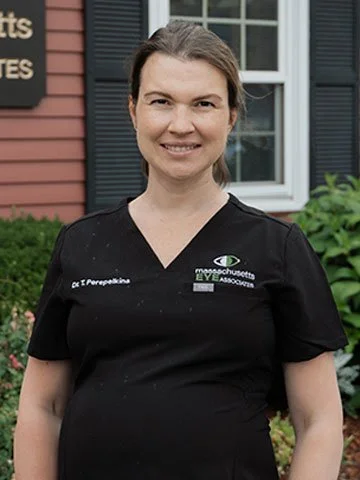 A smiling woman standing outdoors in front of a building with black shutters and green plants, wearing a black medical uniform with the logos of MassEyeAndEar and Dr. Perea-Papadakis.