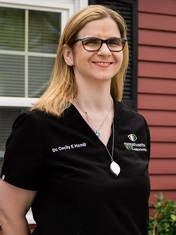 A woman with glasses and blonde hair wearing a black medical uniform with her name and a logo, standing outside a house with red siding and a window.