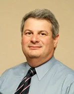 A man with gray hair wearing a light blue dress shirt and a striped tie, smiling at the camera.