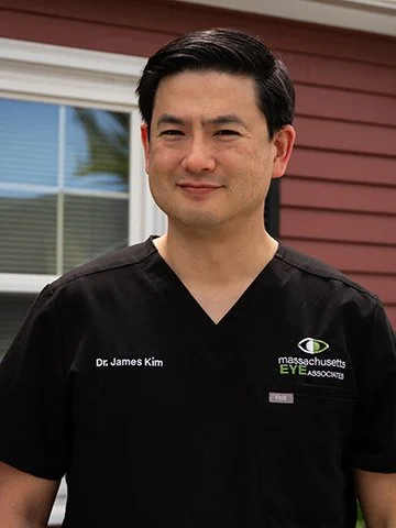 A man wearing a black scrub top with 'Dr. James Kim' and 'Massachusetts Eye Associates' embroidered on it, standing outdoors in front of a house with a window and red siding.