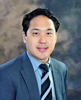 Professional portrait of a young man in a blue suit, blue shirt, and striped tie, with dark hair, against a neutral background.