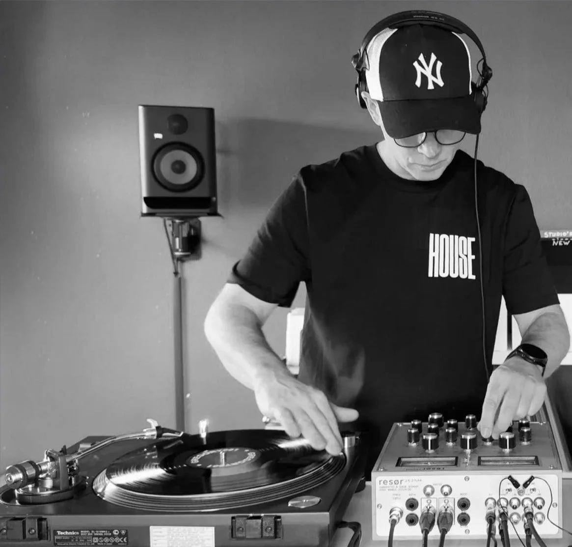 Ein DJ in einem Raum mit Lautsprecher, der Musik auf einem Turntable und einem Mischpult spielt, getragen von einer Person mit Kappe, Brille und einem schwarzen T-Shirt mit weißem Text.