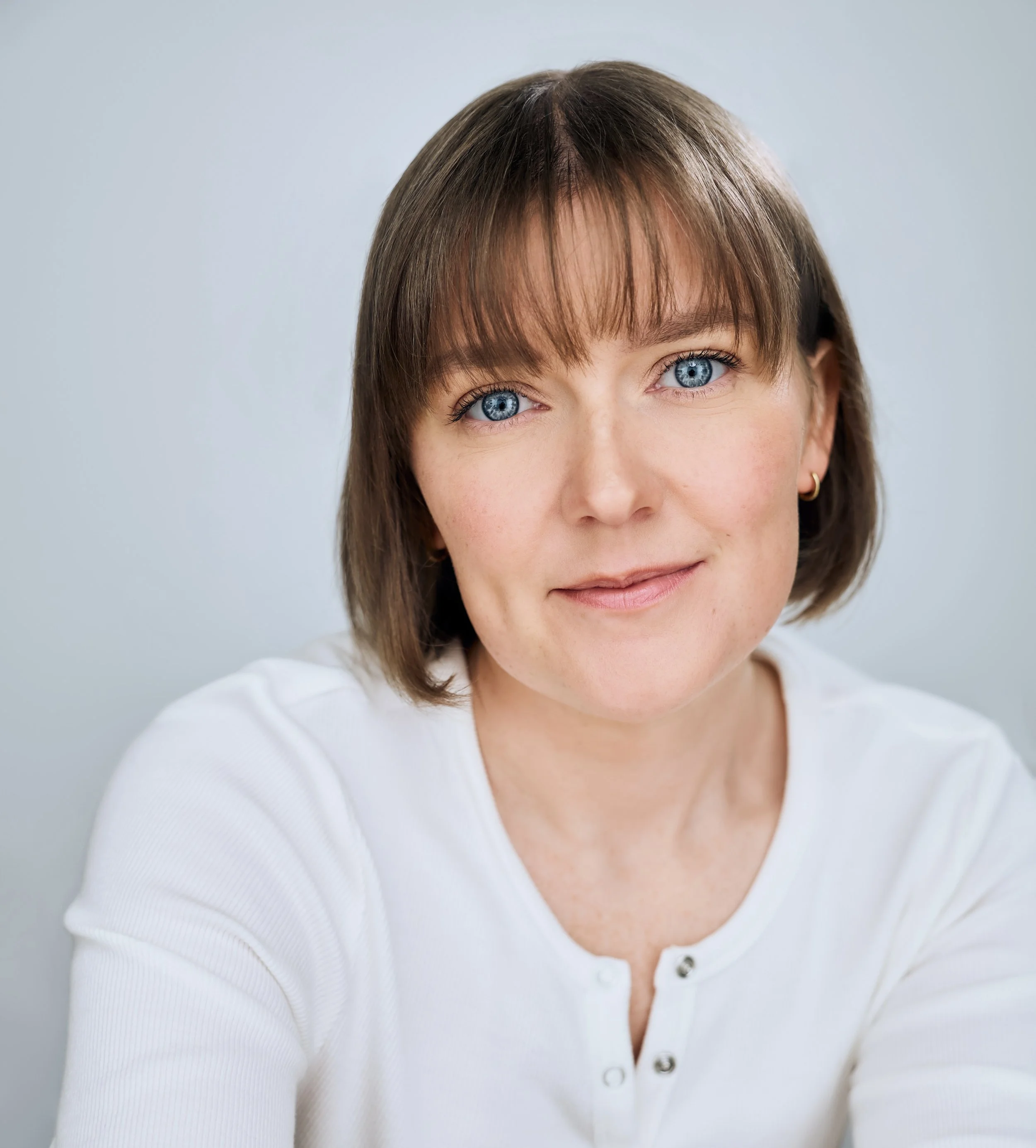 Headshot of actor Meredith Aleigha Wells, who has an ash blonde bob with bangs and striking blue eyes, featuring a warm expression, wearing a white Henley against a light blue background.