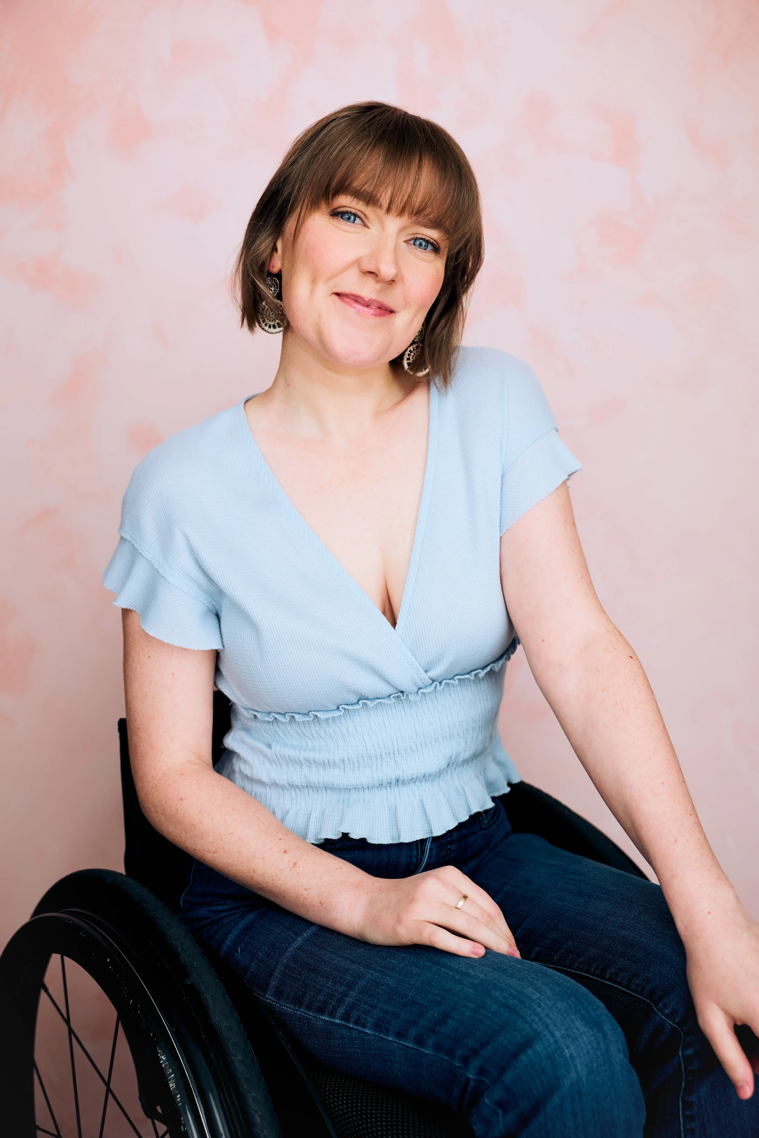 Three-quarter length portrait of actor Meredith Aleigha Wells, who has an ash blonde bob with bangs and expressive blue eyes, featuring an approachable expression, wearing a light blue top and dark wash jeans against a light pink background.