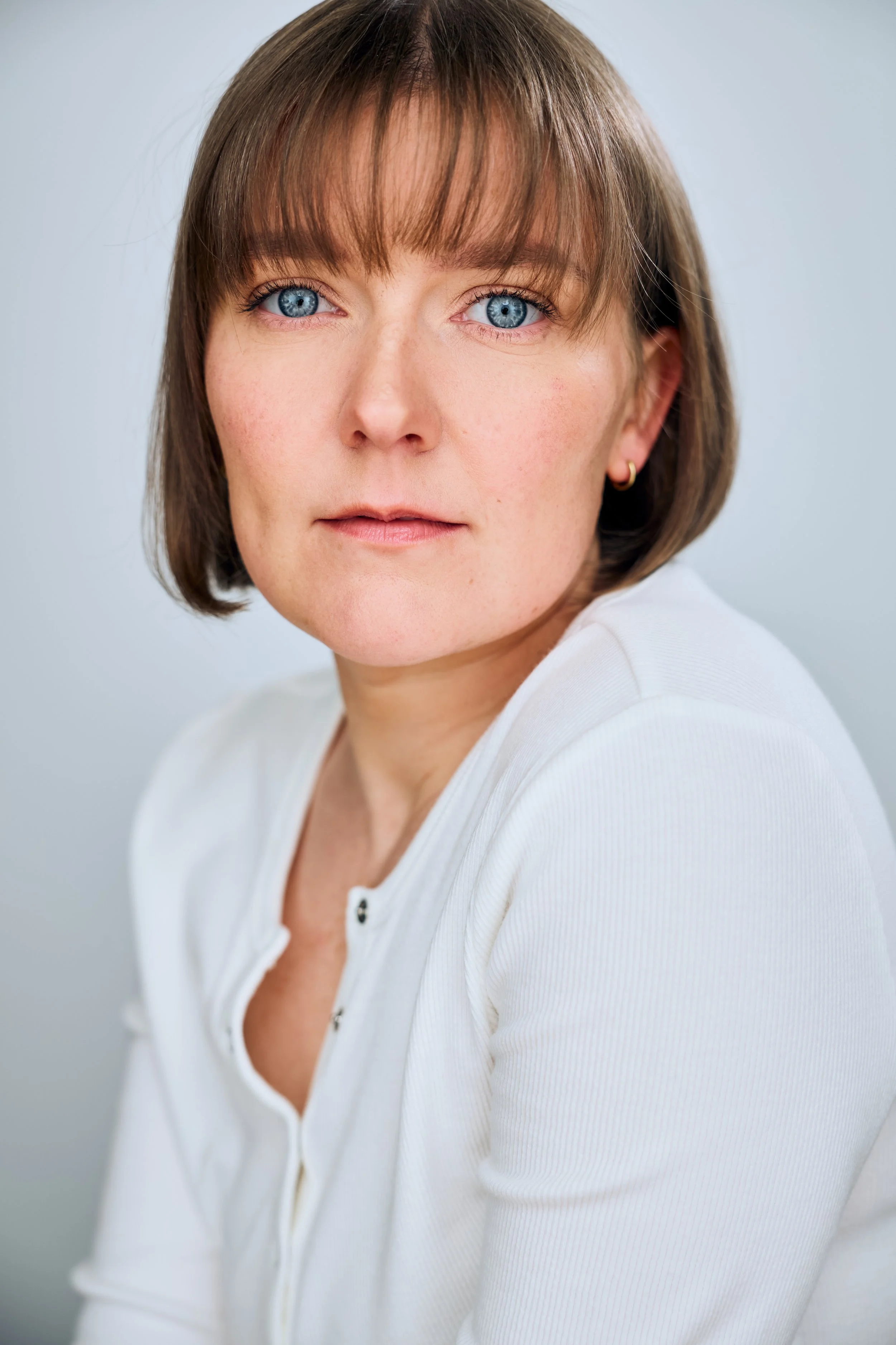 Headshot of actor Meredith Aleigha Wells, who has an ash blonde bob with bangs and striking blue eyes, featuring a piercing expression, wearing a white Henley against a light blue background.
