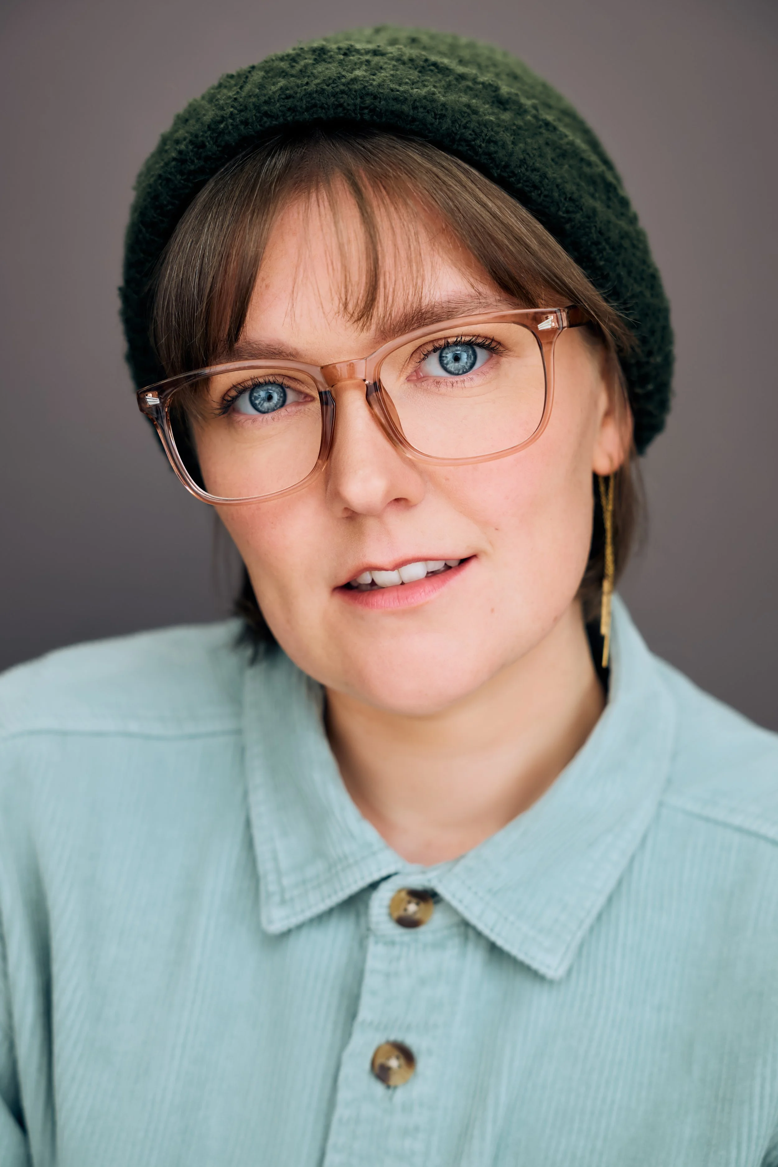 Theatrical Headshot of actor Meredith Aleigha Wells, who has striking blue eyes, featuring an alluring, mid-breath expression with slightly parted lips, wearing a soft aqua corduroy button-up, forest green beanie, and large-framed transparent pink gl
