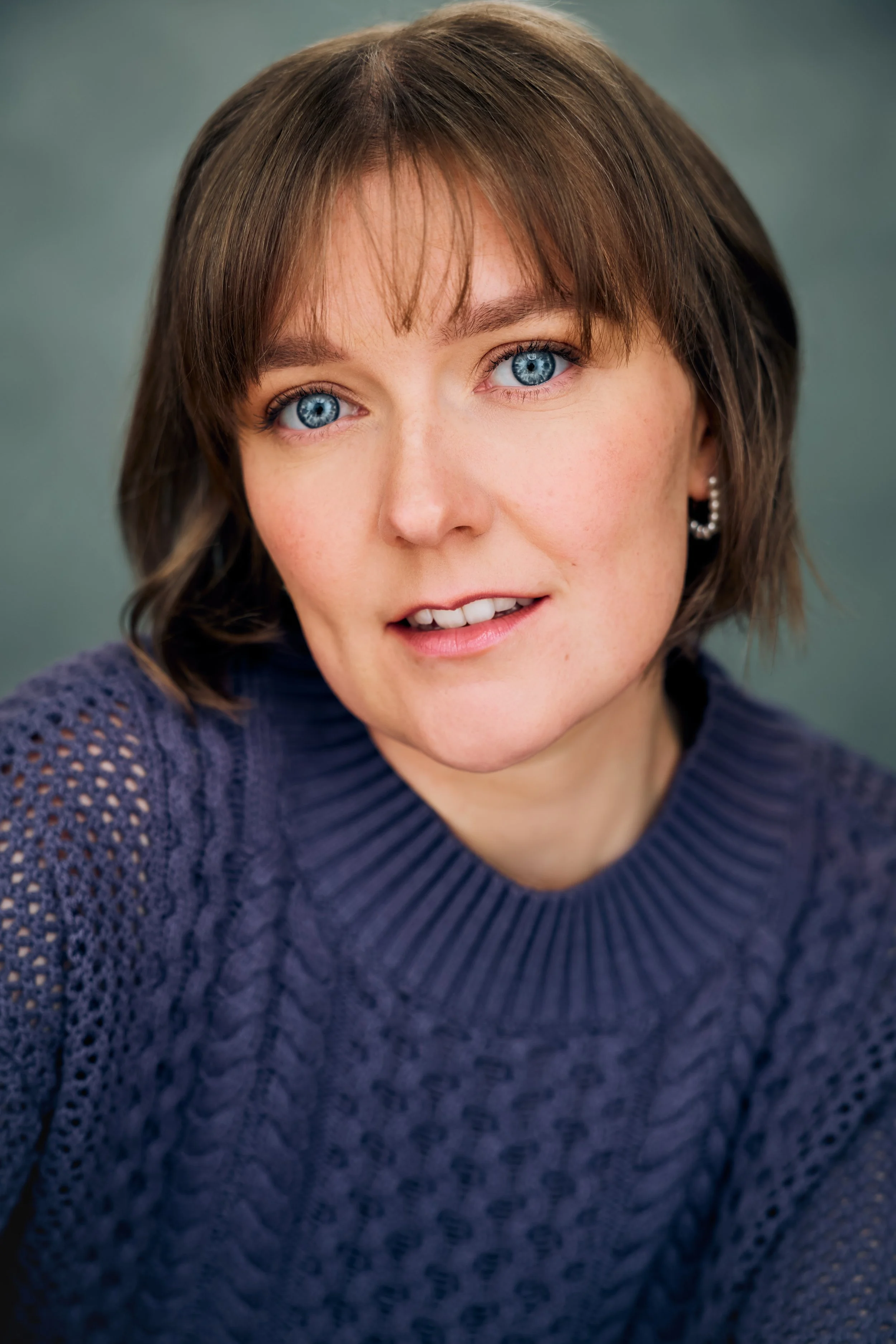 Theatrical Headshot of actor Meredith Aleigha Wells, who has an ash blonde bob with bangs and striking blue eyes, featuring an introspective expression, wearing a chunky indigo sweater against a soft teal background.