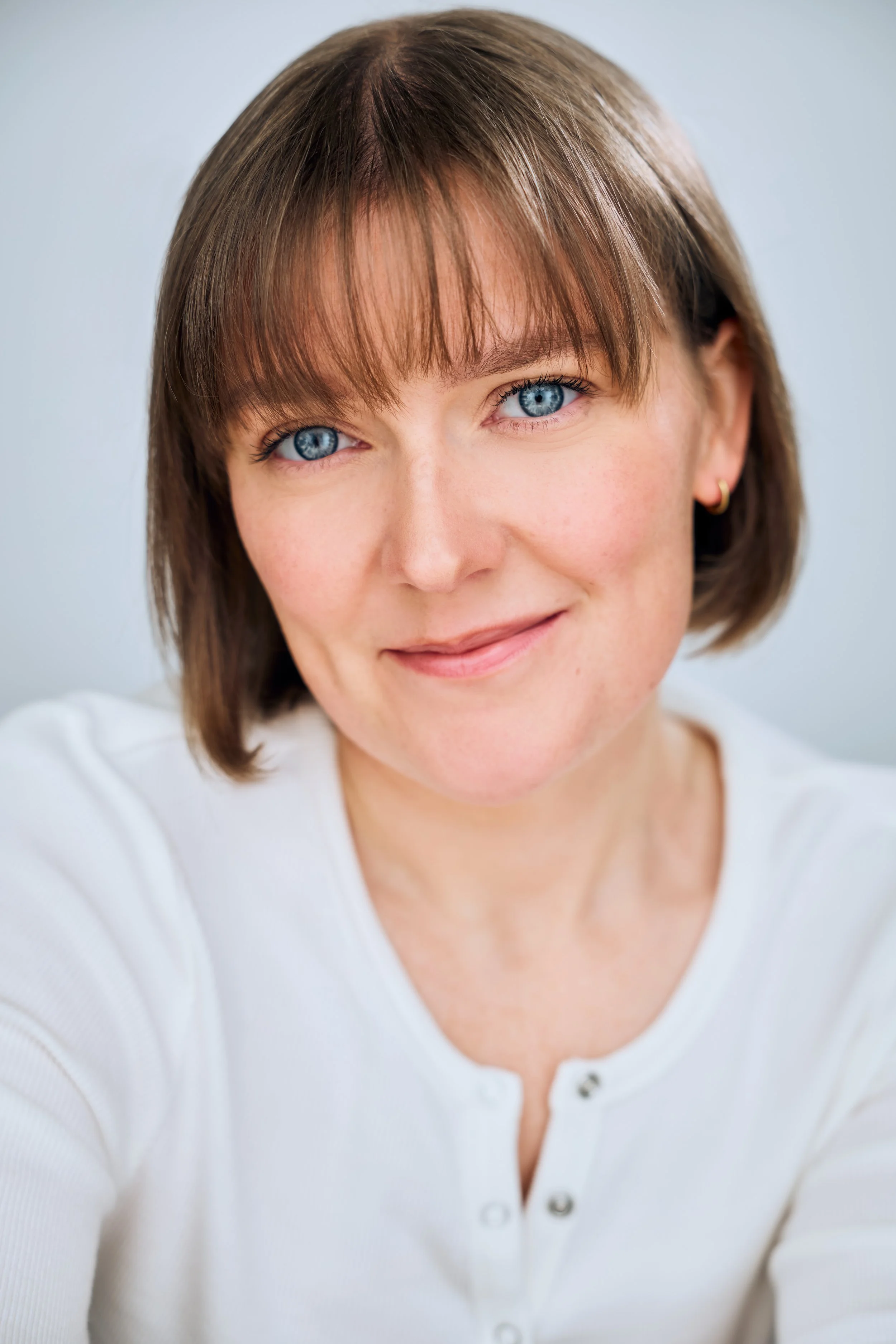 Headshot of actor Meredith Aleigha Wells, who has an ash blonde bob with bangs and expressive blue eyes, featuring a luminous expression, wearing a white Henley against a light blue background.