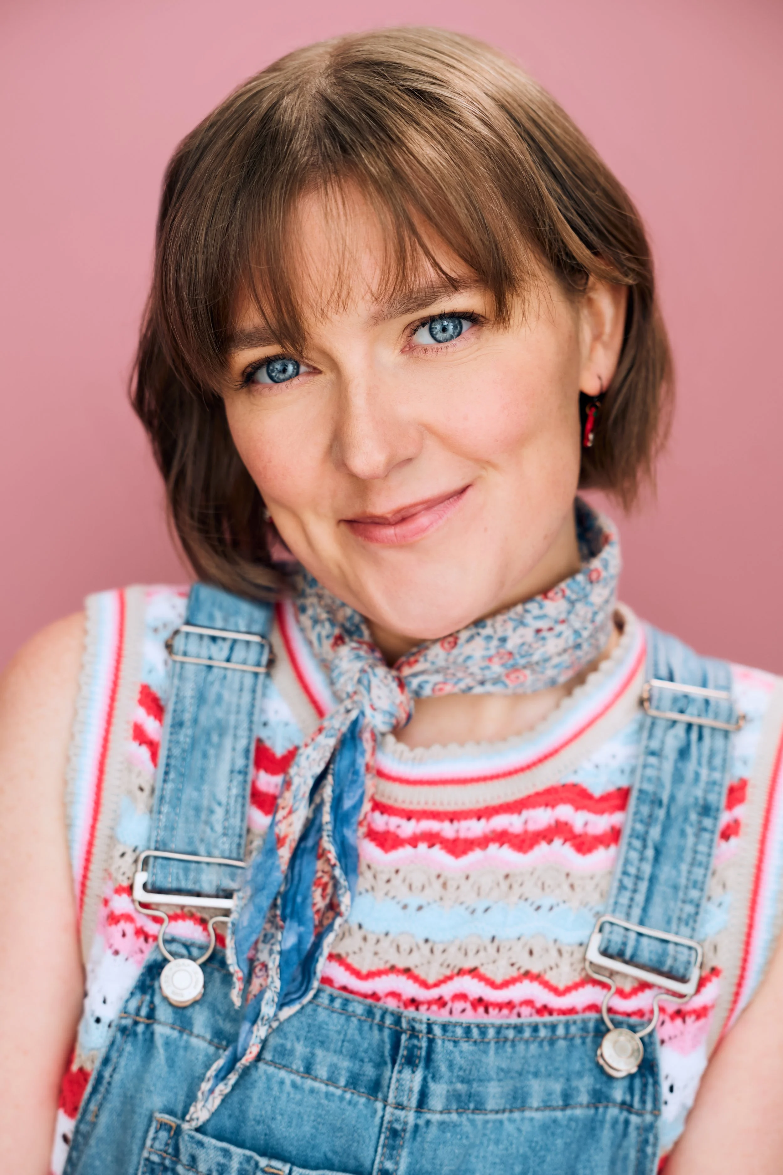 Commercial Headshot of actor Meredith Aleigha Wells, who has an ash blonde bob with bangs and expressive blue eyes, featuring a mischievous expression, wearing denim overalls and a bright patterned sleeveless sweater and neckerchief against a bright 