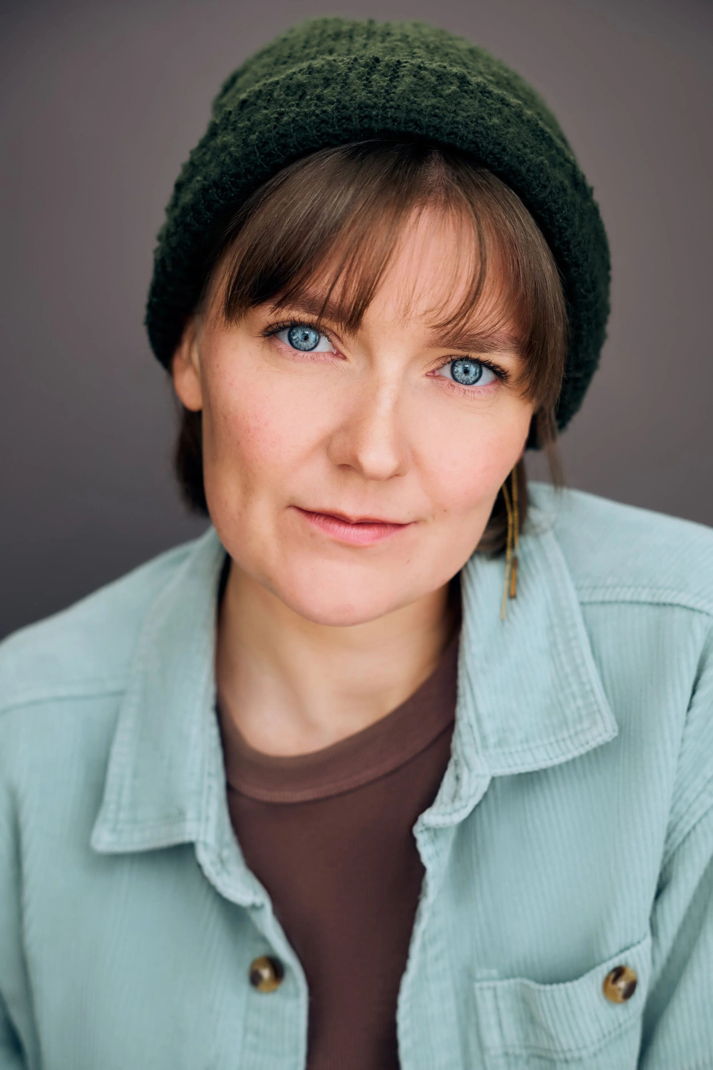 Theatrical Headshot of actor Meredith Aleigha Wells, who has striking blue eyes, featuring a grounded expression, wearing a soft aqua corduroy button-up over a taupe T-shirt, and a forest green beanie.
