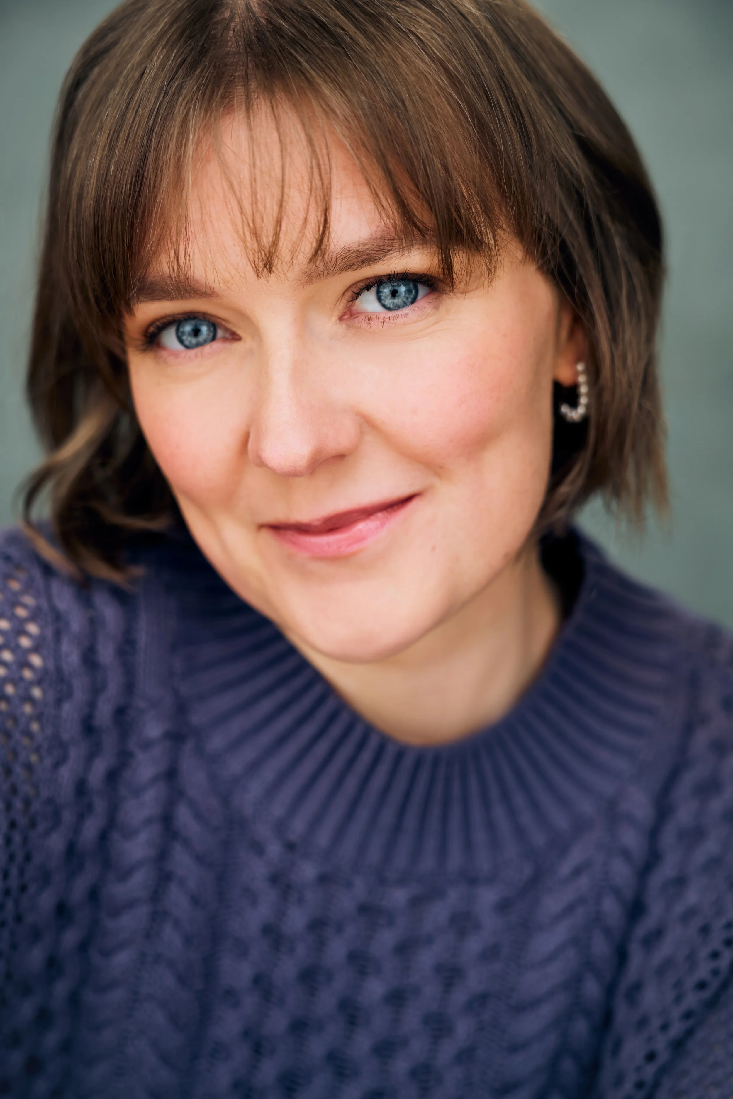 Headshot of actor Meredith Aleigha Wells, who has an ash blonde bob with bangs and striking blue eyes, featuring a warm expression, an indigo sweater against a soft teal background.