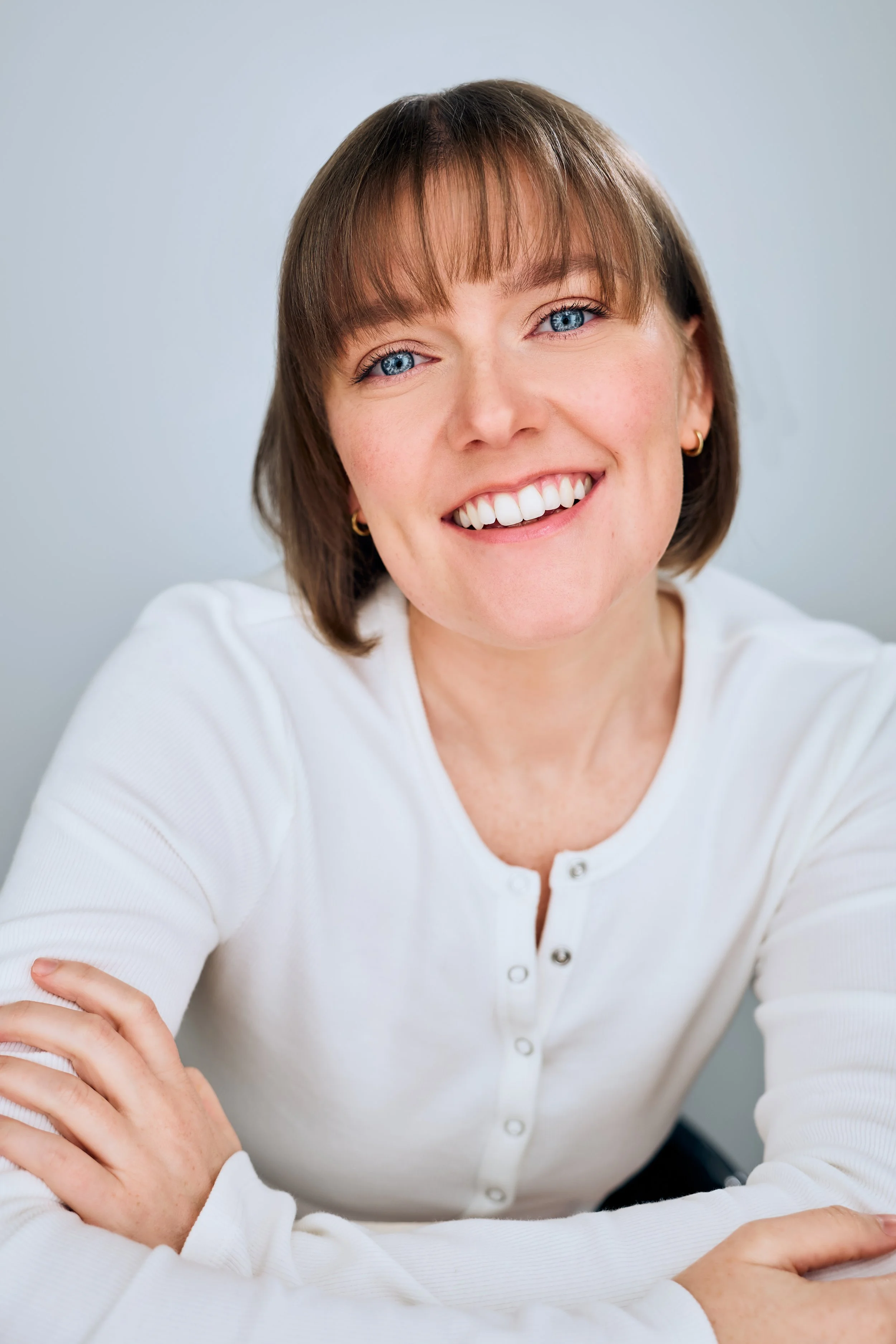 Headshot of actor Meredith Aleigha Wells, who has an ash blonde bob with bangs and expressive blue eyes, featuring an effervescent expression, wearing a white Henley against a light blue background.
