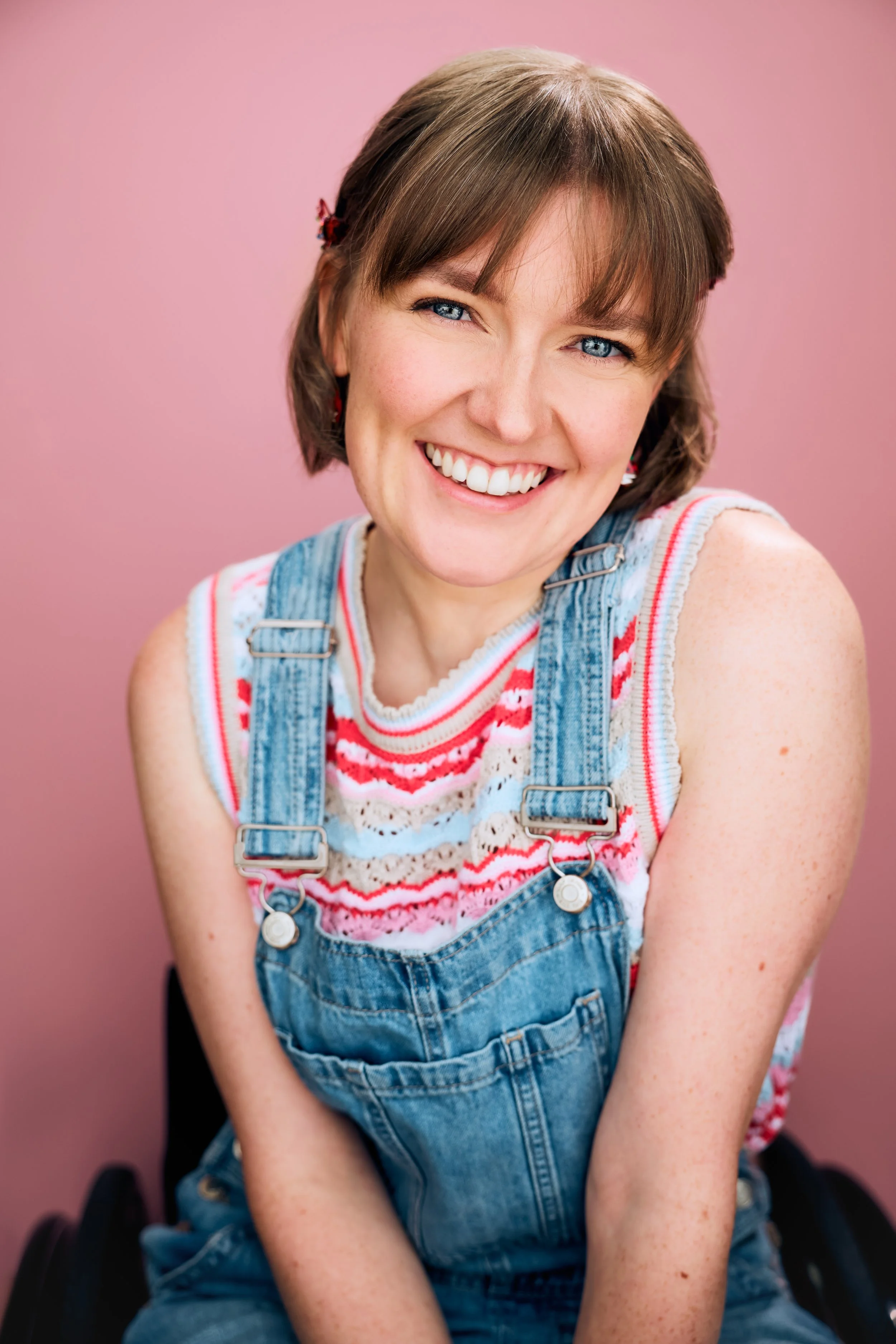 Three-Quarter Length Portrait of actor Meredith Aleigha Wells, who has an ash blonde bob with bangs and expressive blue eyes, featuring a bubbly expression, wearing denim overalls and a bright patterned sleeveless sweater and neckerchief against a br