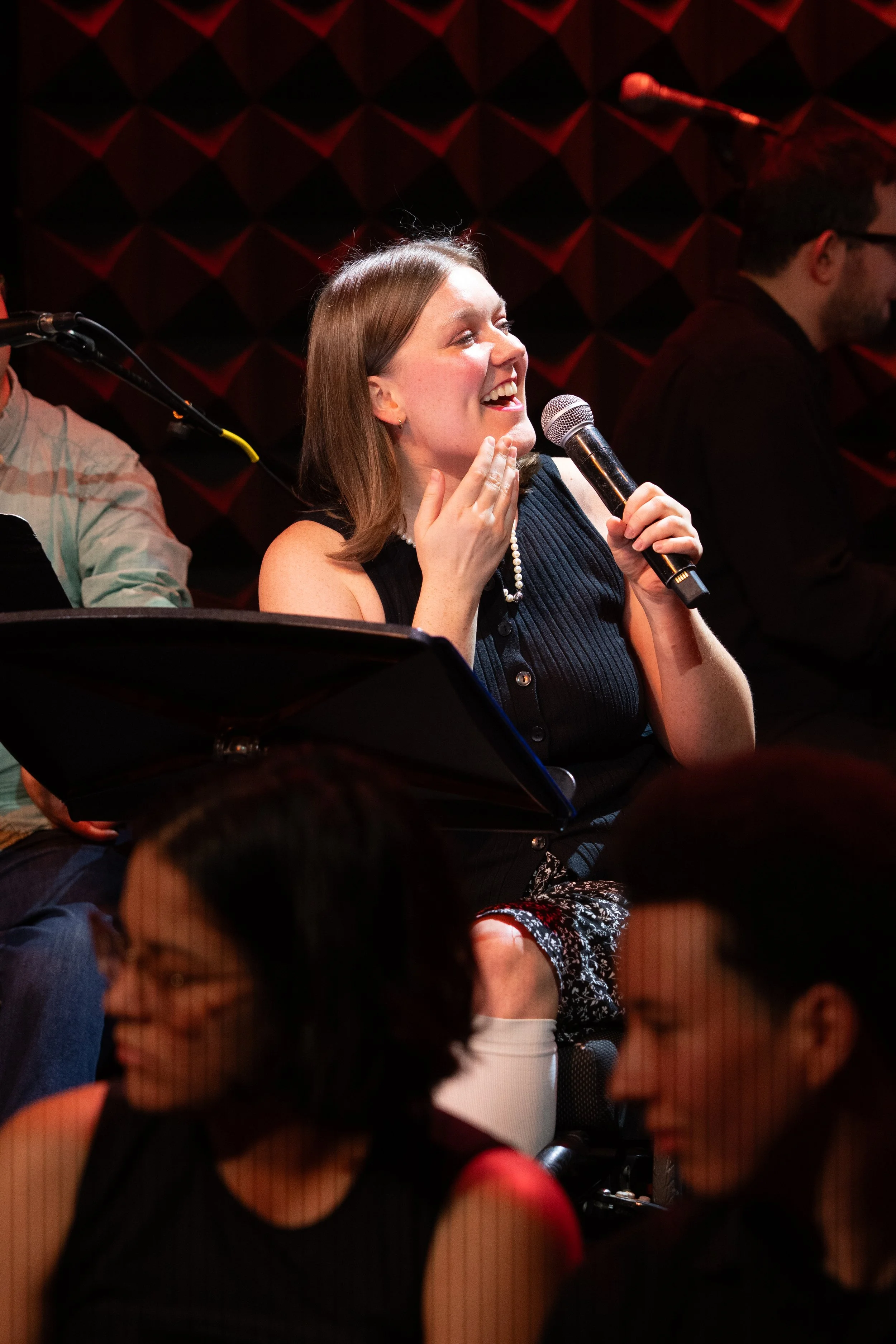 A white wheelchair user with shoulder length brown hair sings into a microphone and signs thank you with the other hand. two asl interpreters sit in the foreground and the Joe’s Pub stage is in the background.
