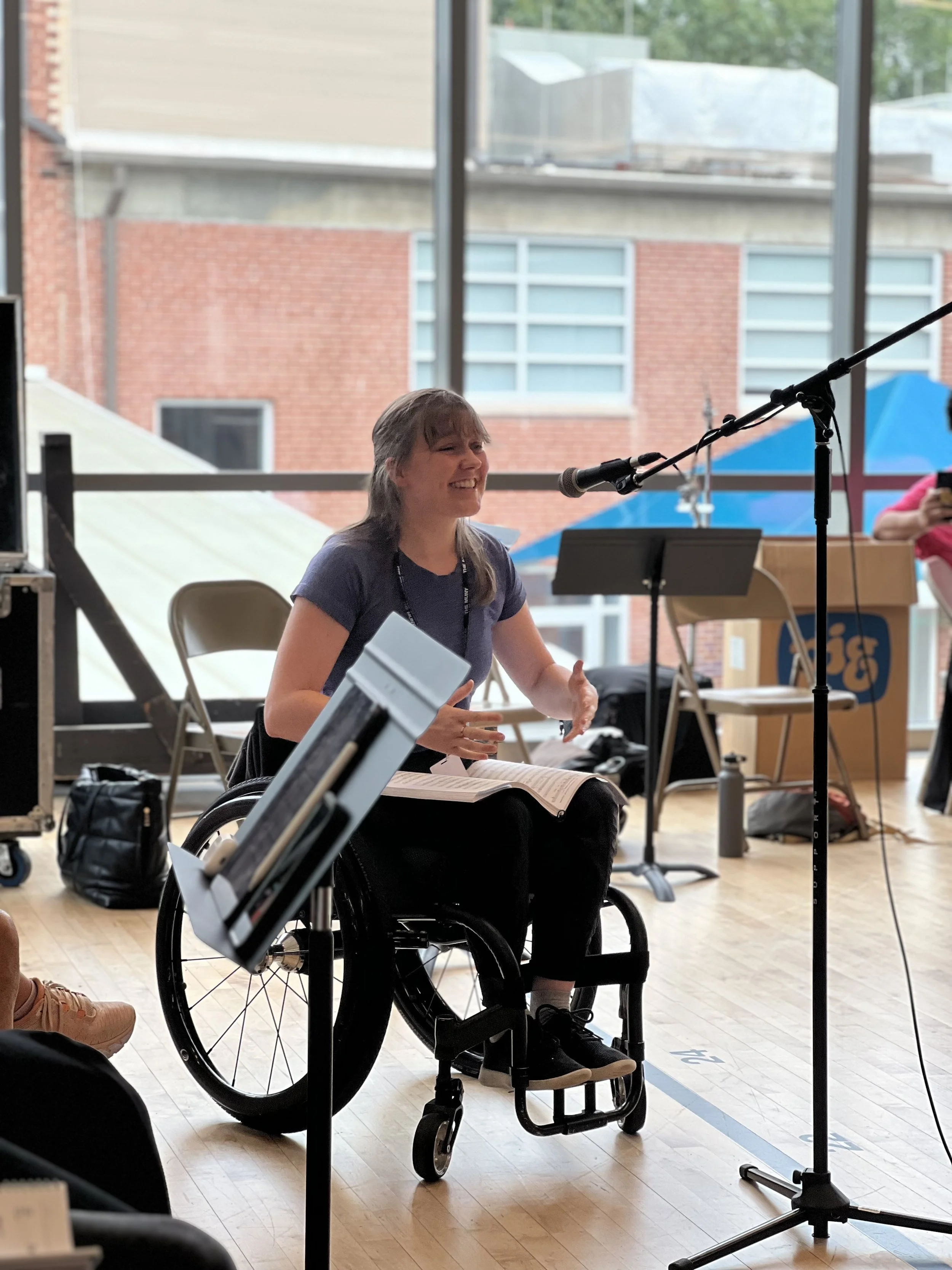candid photo of musical theatre artist Meredith Aleigha Wells sitting in their wheelchair singing at a microphone, inside a brightly lit rehearsal room with large floor to ceiling windows