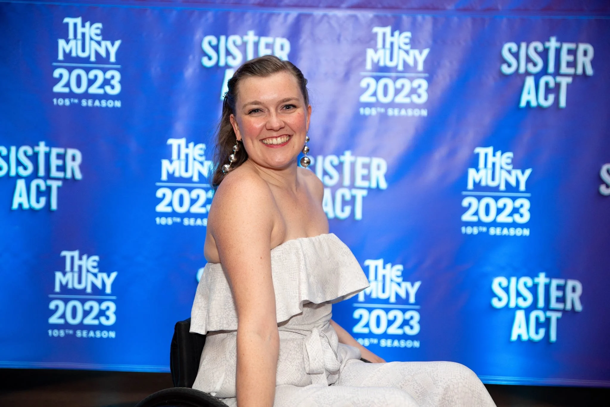 Musical Theatre Artist Meredith Aleigha Wells, a wheelchair user with fair skin and an ash blonde hair, features a radiant smile. They wear a white jumpsuit with dangling disco ball earrings, posing in front of a purple step-and-repeat backdrop