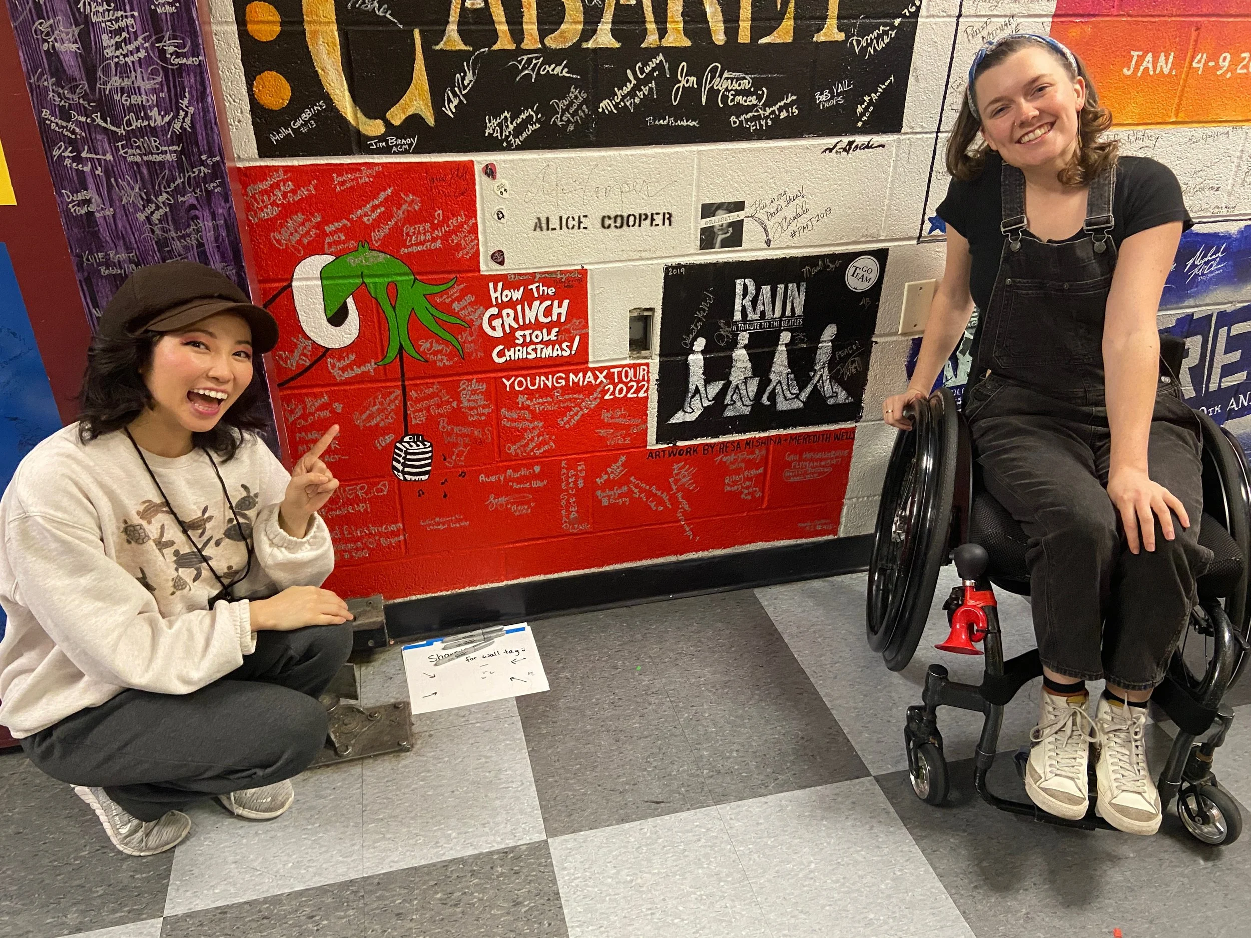 Actor Meredith Aleigha Wells using a wheelchair backstage at the Orpheum Theatre with co-star Resa by their Grinch wall tag painting.
