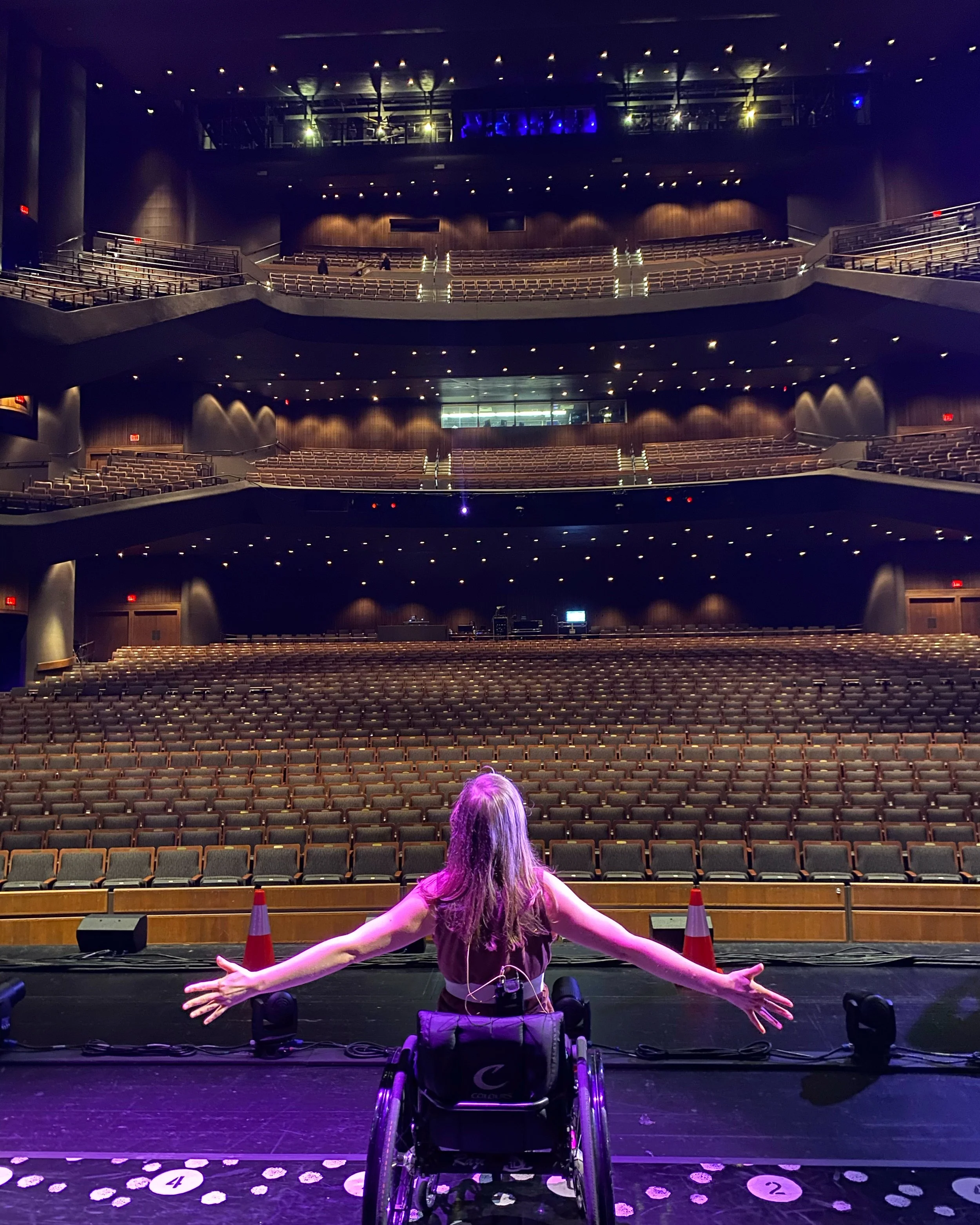 Performer Meredith Aleigha Wells sitting in their wheelchair with their back to the camera and arms outstretched looking out into the house of Bass Concert Hall in purple/pink lighting.