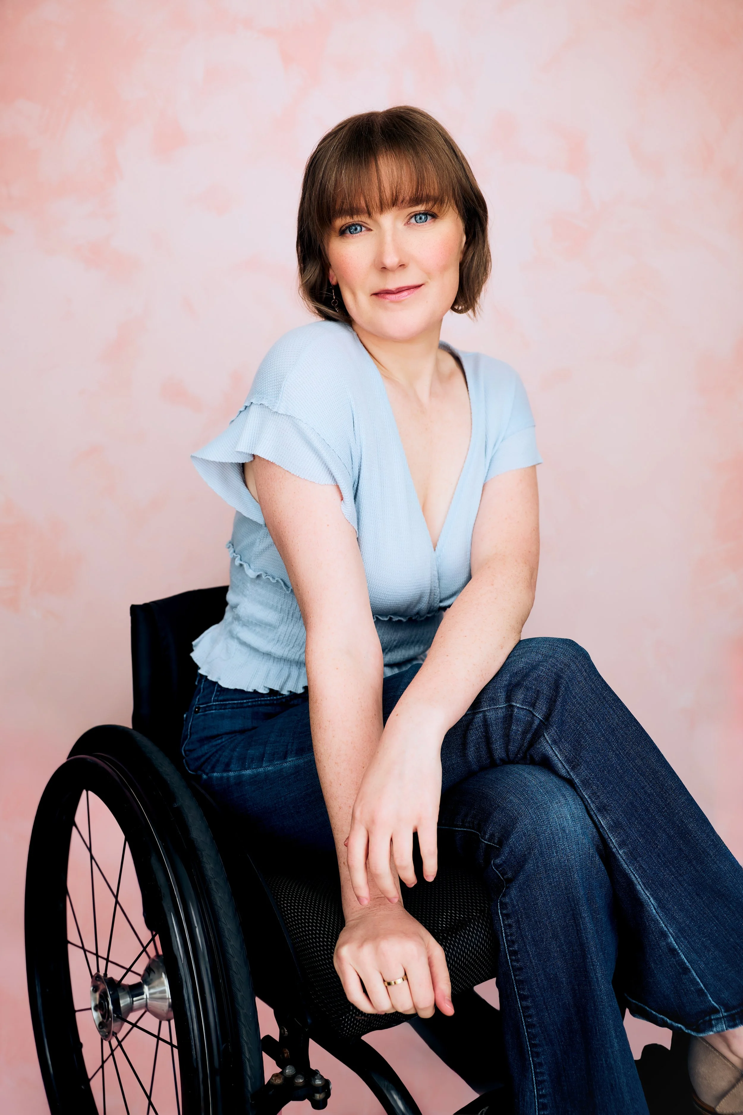 Three-quarter length portrait of actor Meredith Aleigha Wells, who has an ash blonde bob with bangs and striking blue eyes, featuring a tranquil expression, wearing a light blue top and dark wash jeans against a light pink background.
