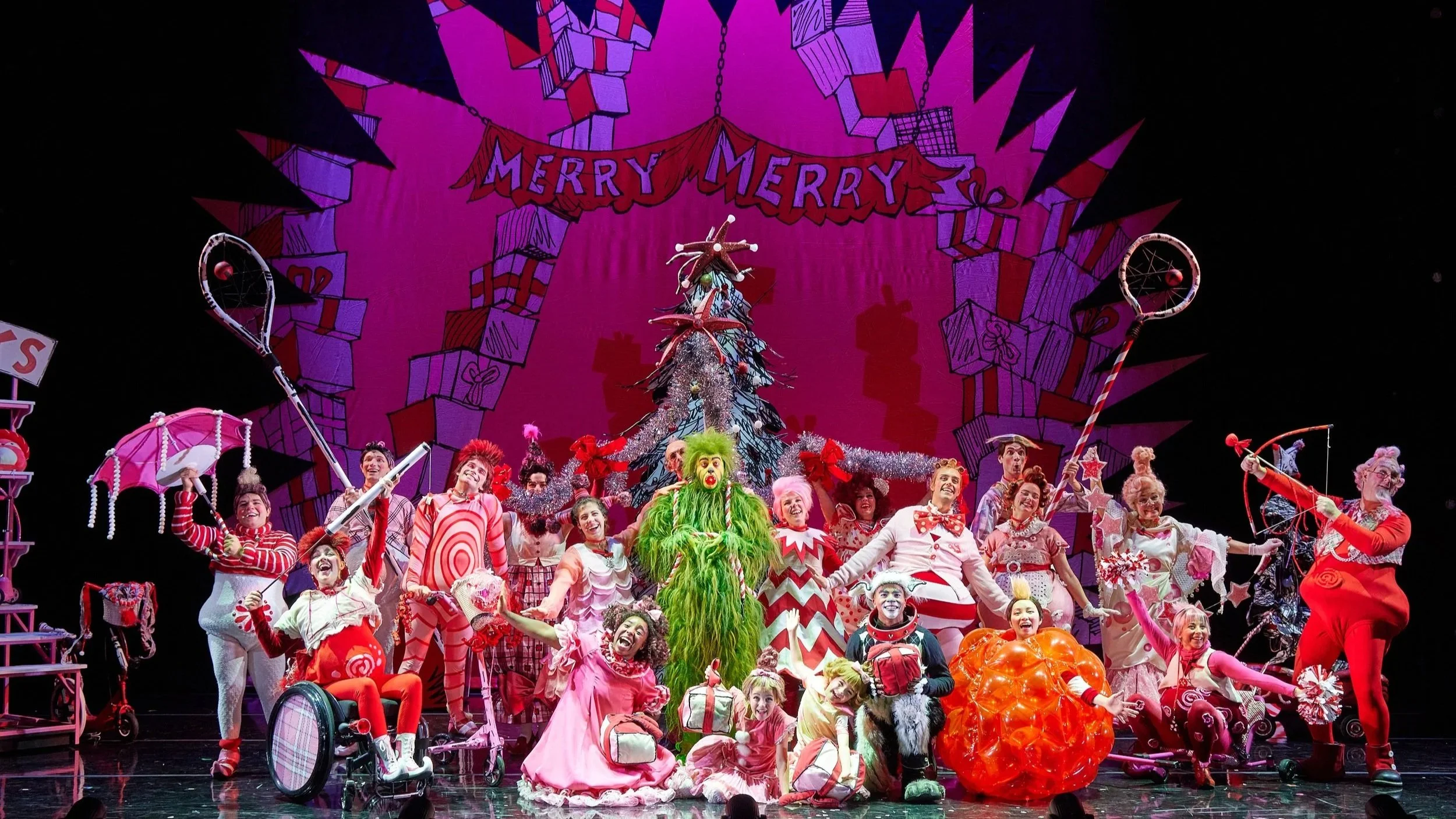 Production still from the Broadway National Tour of Dr. Seuss'  How the Grinch Stole Christmas! the musical, featuring the full cast looking exuberant, posing at the end of a musical number in front of a red, pink, white, and black backdrop