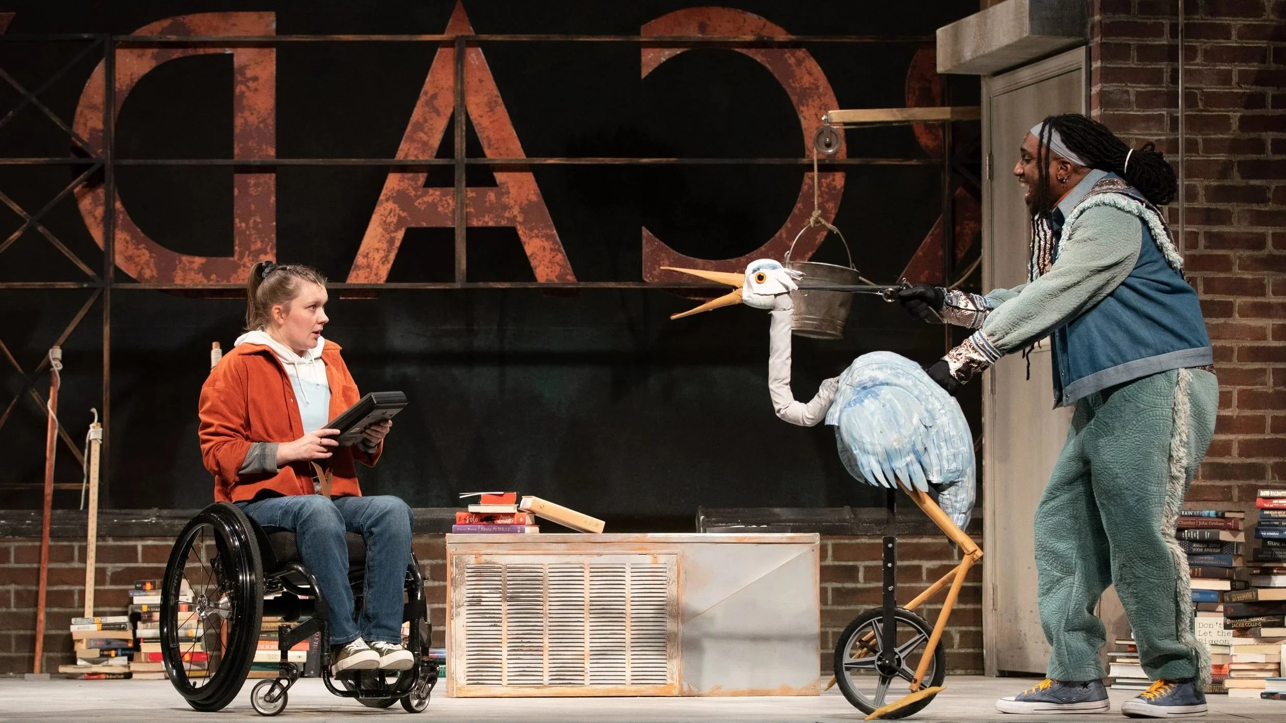 Musical Theatre production still from Through the Sunken Lands at The Kennedy Center featuring actor Meredith Aleigha Wells as Artemis, looking bewildered while staring a blue heron puppet on a rooftop setting.