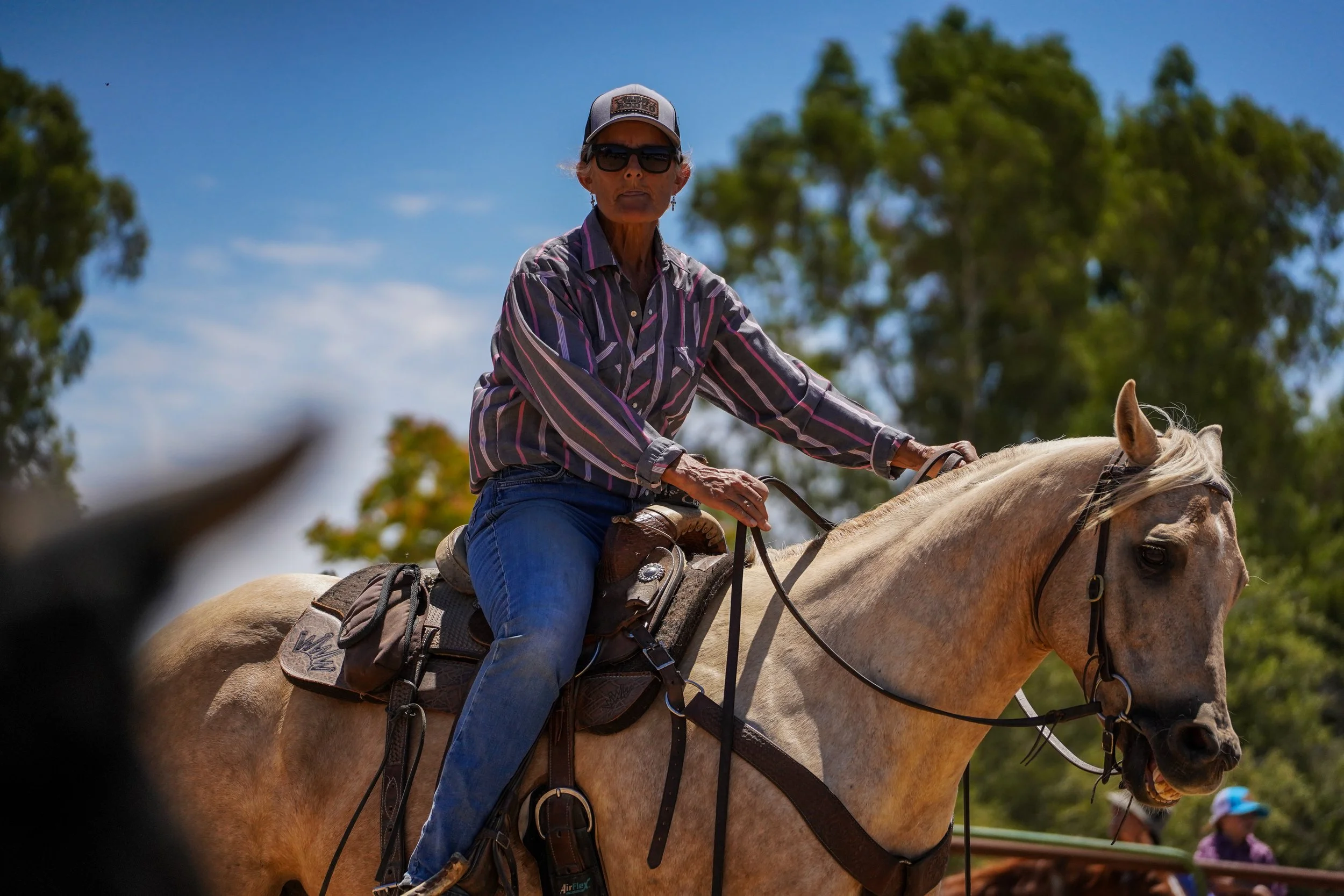 A woman wearing sunglasses, a cap, and a purple striped shirt riding a light-colored horse outdoors. There are trees and a blue sky in the background.