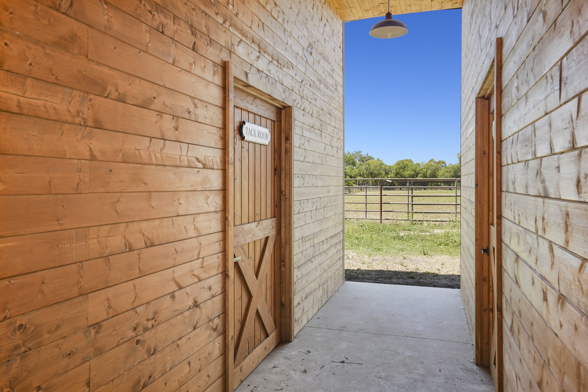 View of a barn stall doorway labeled "Tack Room" with wooden walls and a gate outside showing trees and a clear blue sky.