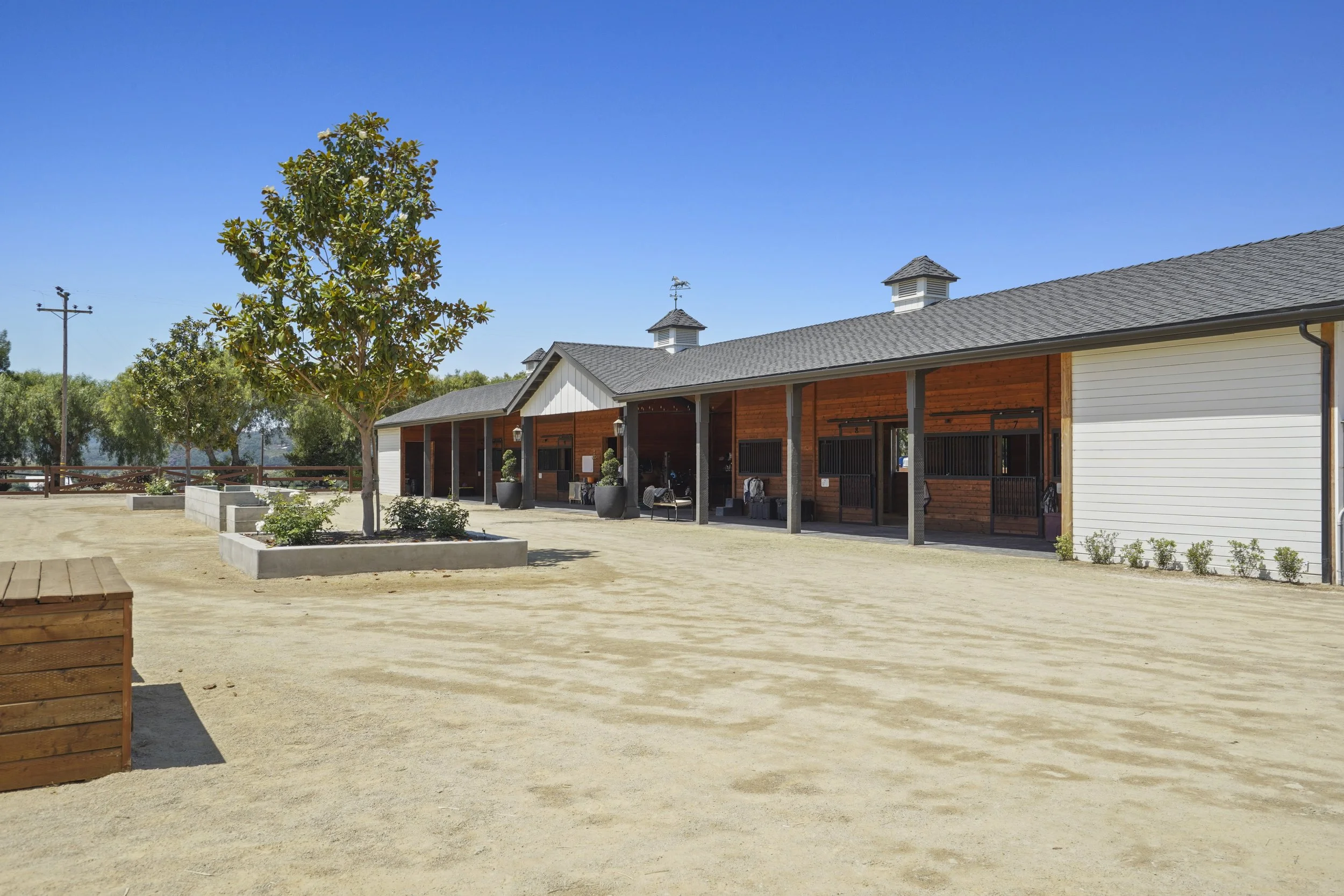 A building with a gray shingled roof and white and wooden siding. There is a tree in front and a dirt area in the foreground. The sky is clear and blue.